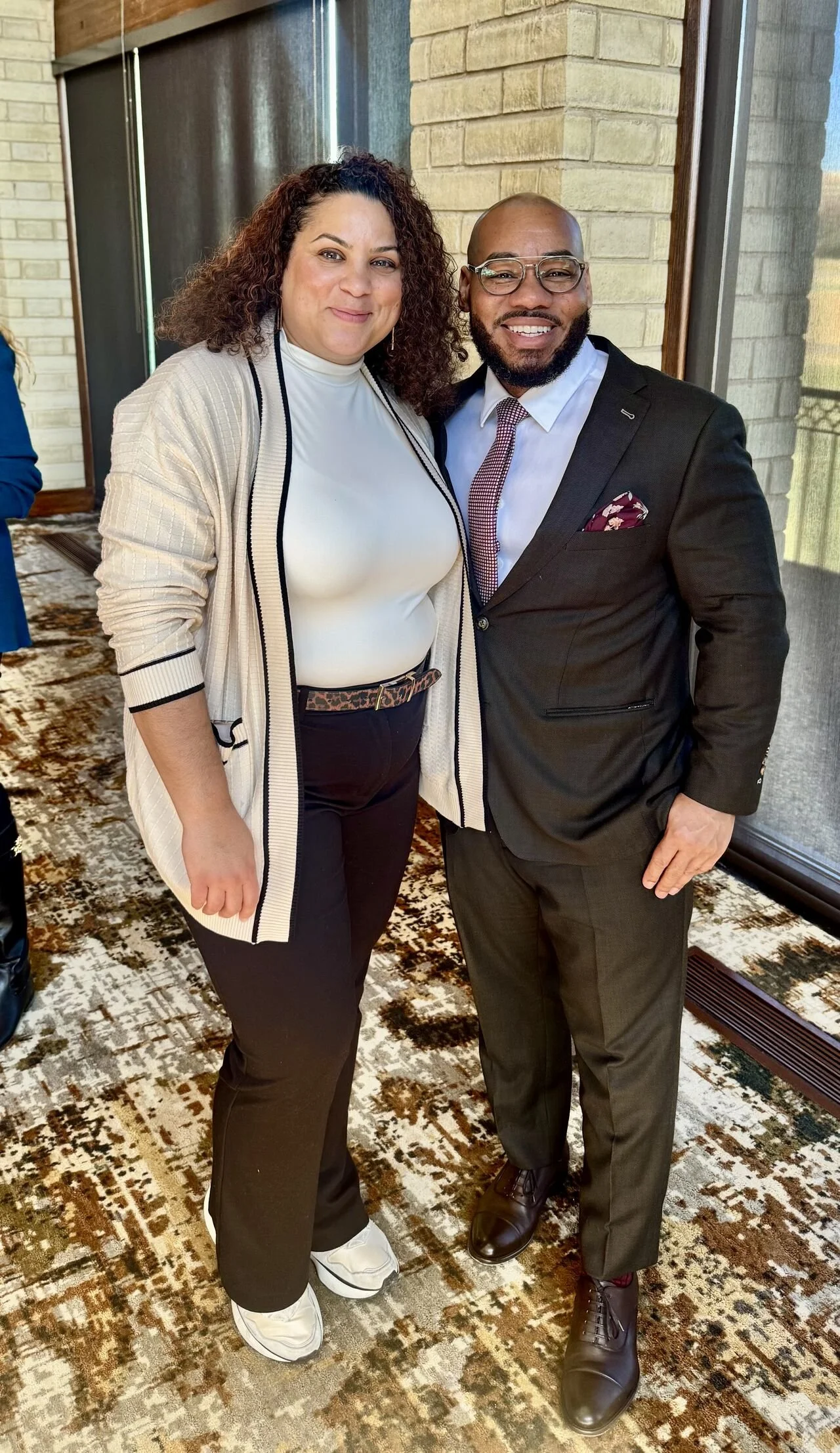 A woman and man standing together indoors, smiling at the camera. The woman has curly hair, is wearing a white turtleneck and black pants, and the man has a beard, is wearing glasses, a suit, and a tie.