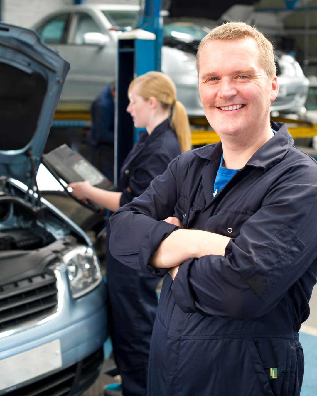 A smiling male auto mechanic standing with arms crossed in a garage, with a woman working on a car engine in the background, and other vehicles on car lifts.