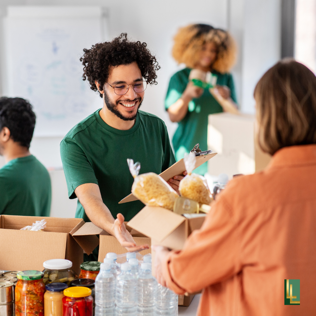 A smiling man in glasses handing a box of food to a woman at a community service or food distribution event, with other volunteers and supplies in the background.
