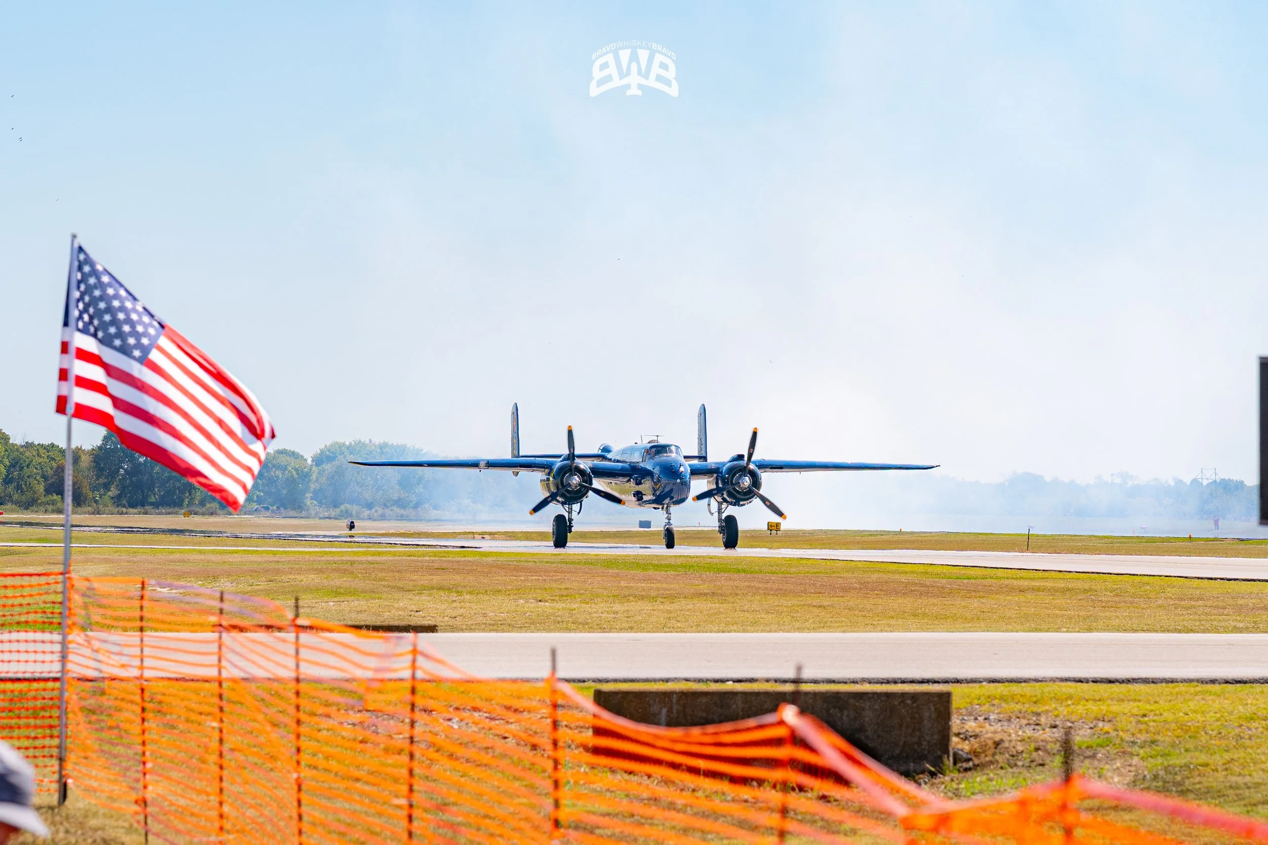 A vintage military aircraft taxiing on the runway with an American flag in the foreground and orange safety fencing on the ground.