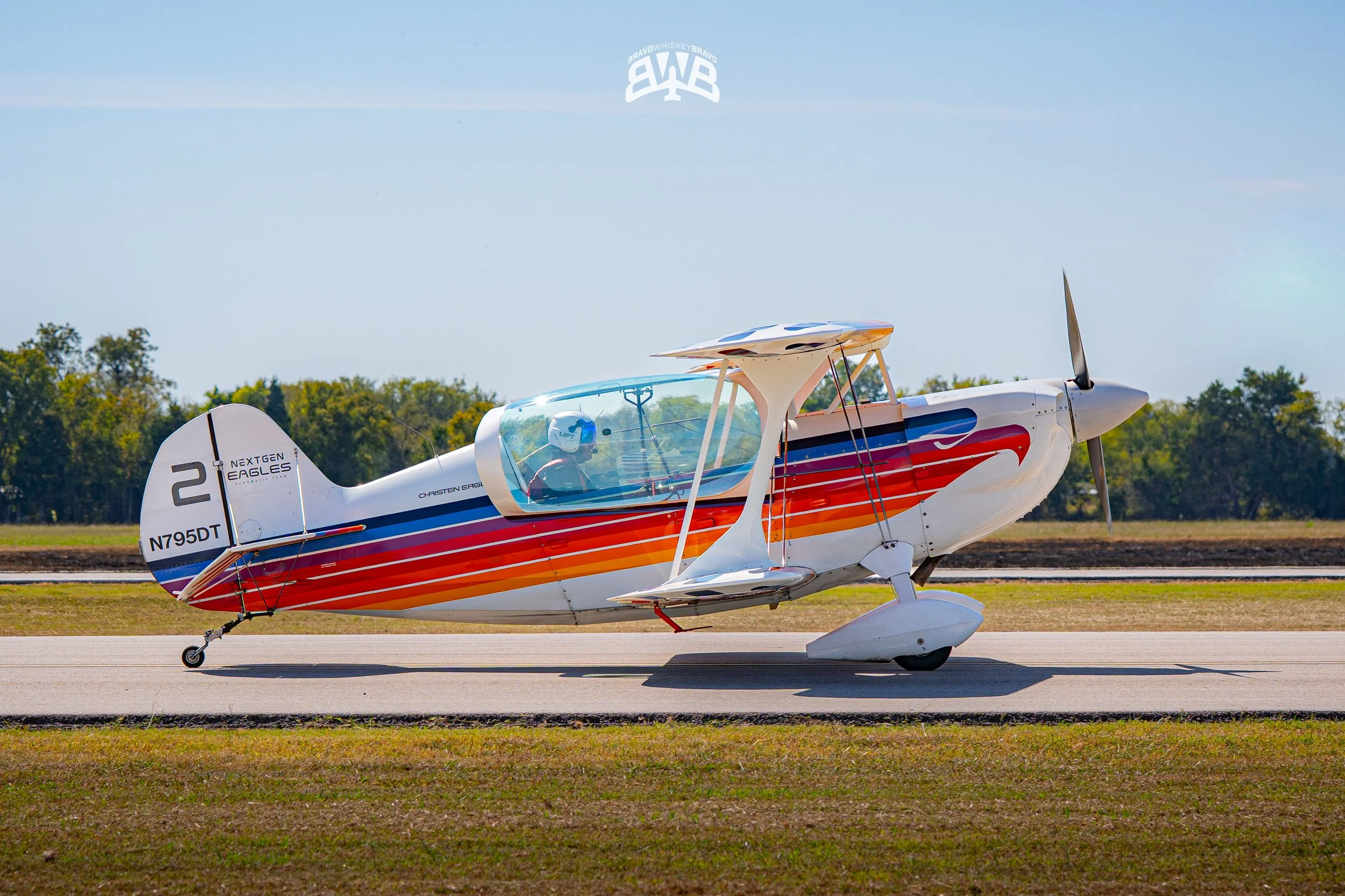 A colorful single-engine aircraft parked on a runway, with a pilot inside wearing a helmet, and trees in the background.