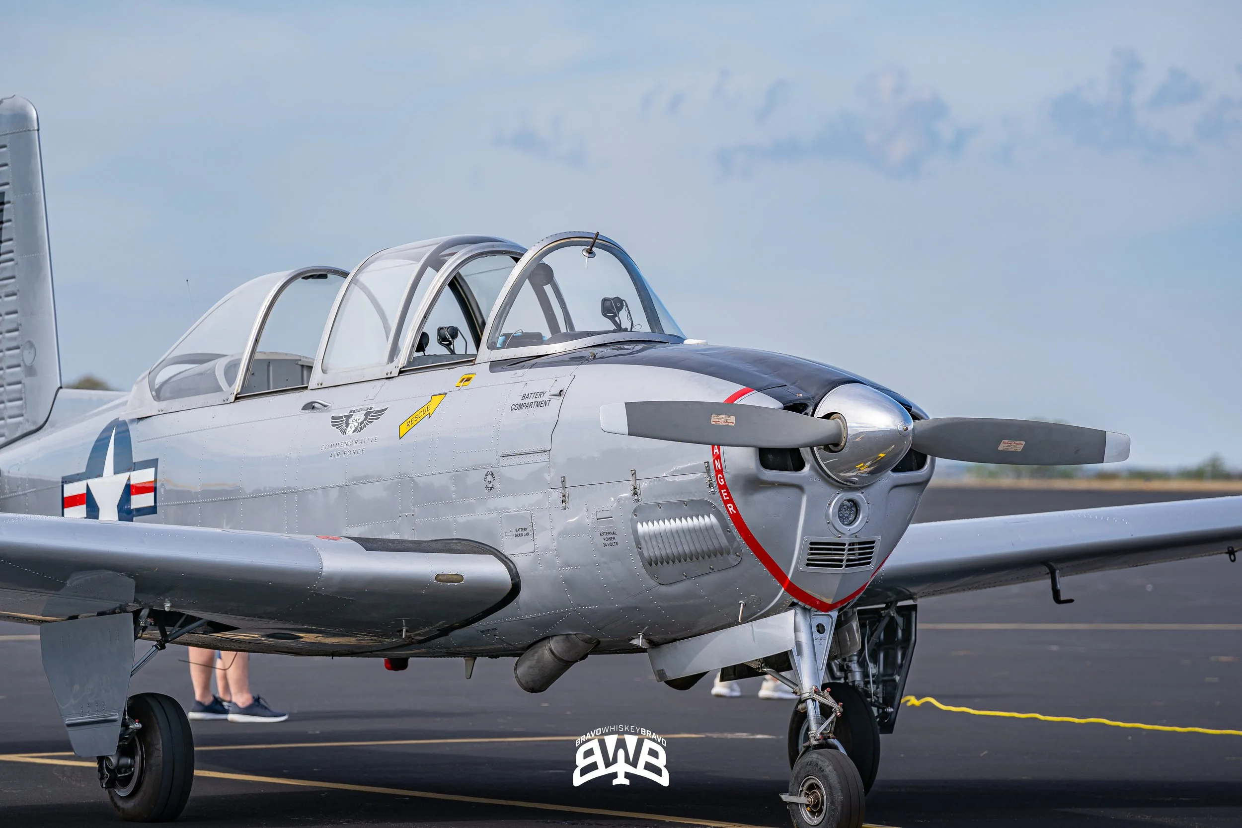 A vintage silver military aircraft with a propeller, parked on a tarmac under a blue sky, with a person standing in the background.
