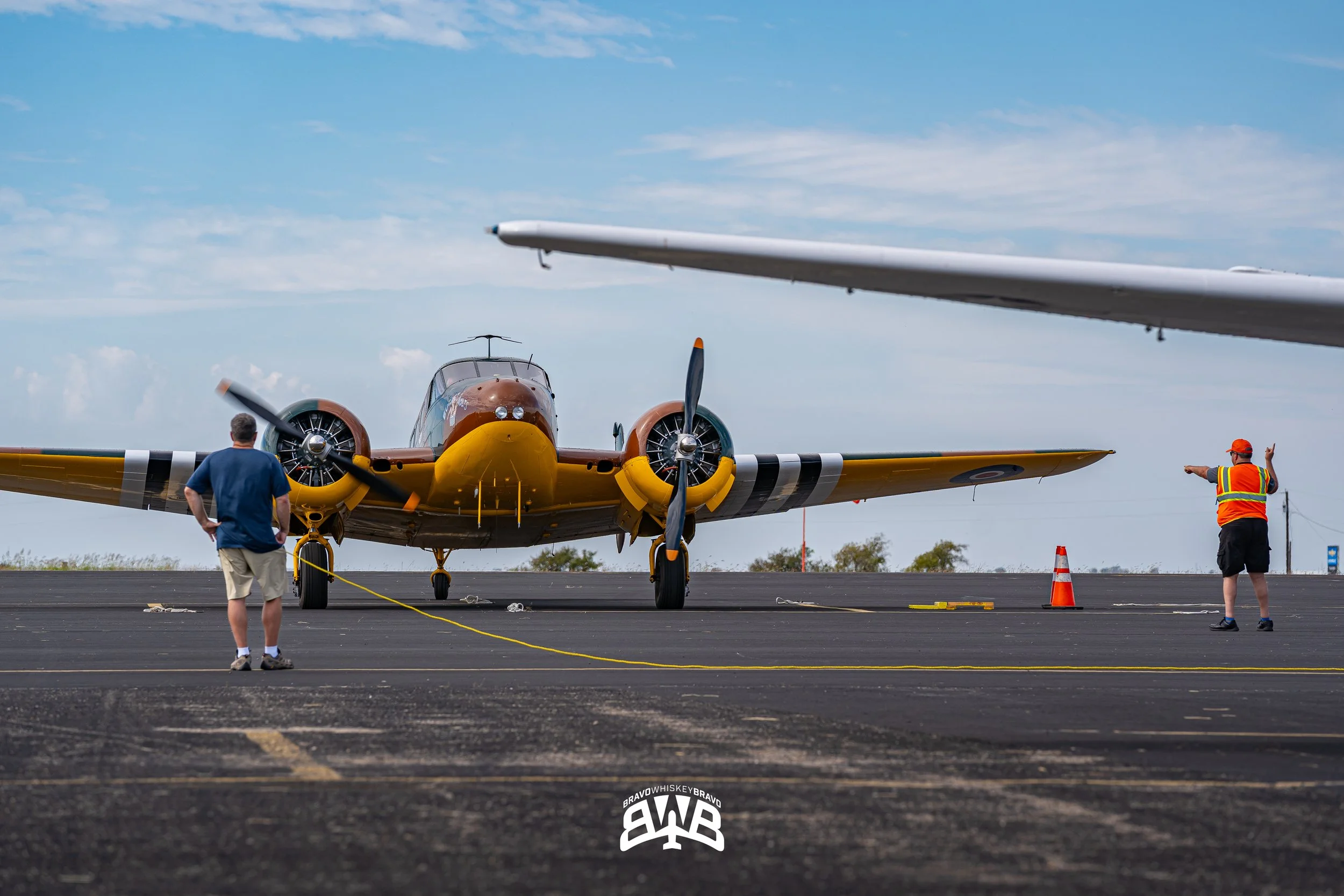 A vintage yellow and brown aircraft is on the tarmac with two people near it, one in a blue shirt and beige shorts holding a yellow rope, and another in a safety vest and orange hat signaling. The sky is partly cloudy.