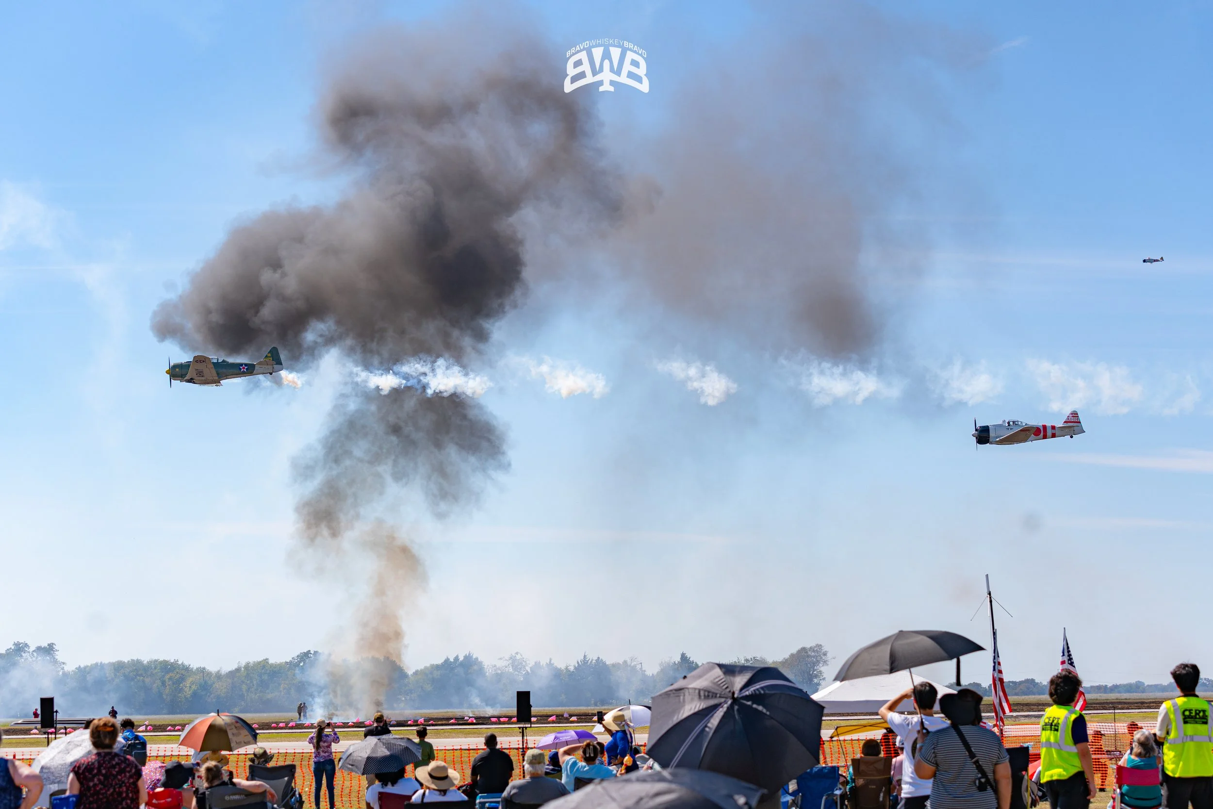 Vintage aircraft flying over an airshow, with smoke and fire in the sky, and spectators watching below.