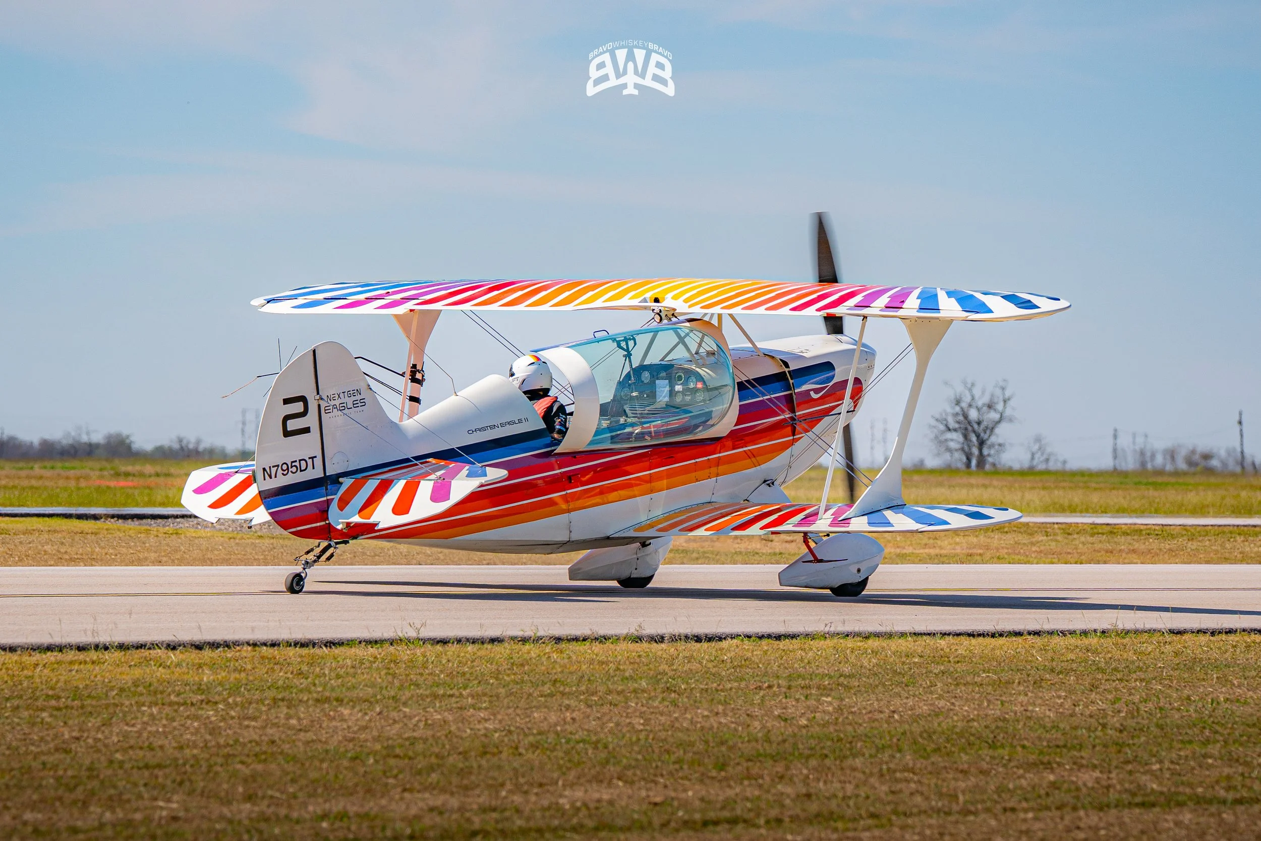 Colorful biplane on runway with rainbow-colored wings and tail, cockpit with pilot in helmet, open sky in background.