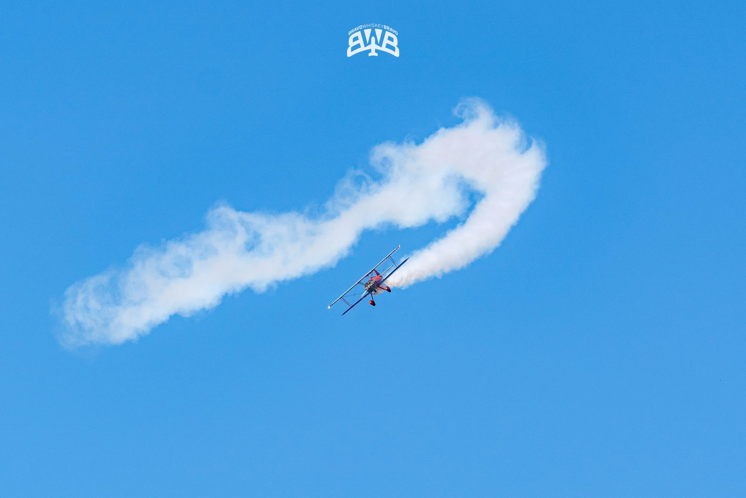 A red biplane performing an aerobatic stunt in the sky, leaving a trail of smoke behind.