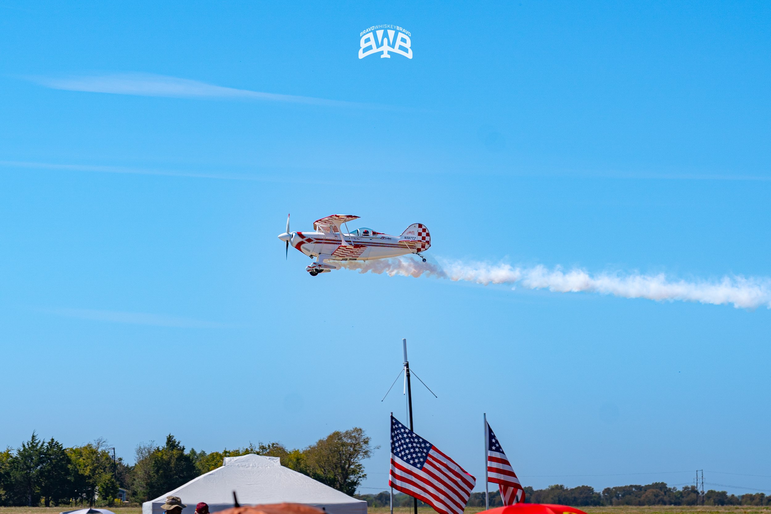 A small aircraft performing a stunt in the sky with a trail of white smoke, American flags on the ground, and a clear blue sky.