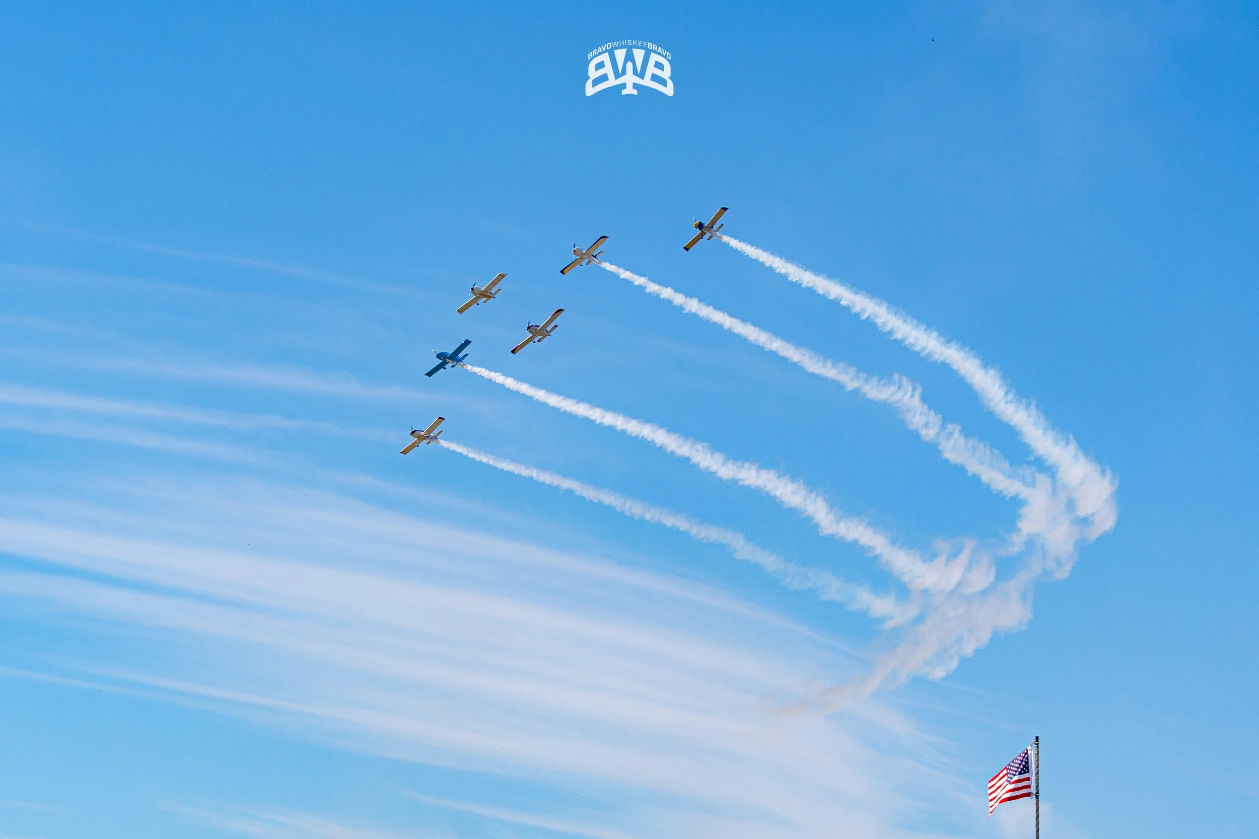 Six small airplanes flying in a formation in the sky, with white smoke trails behind them, and an American flag waving at the bottom right corner.
