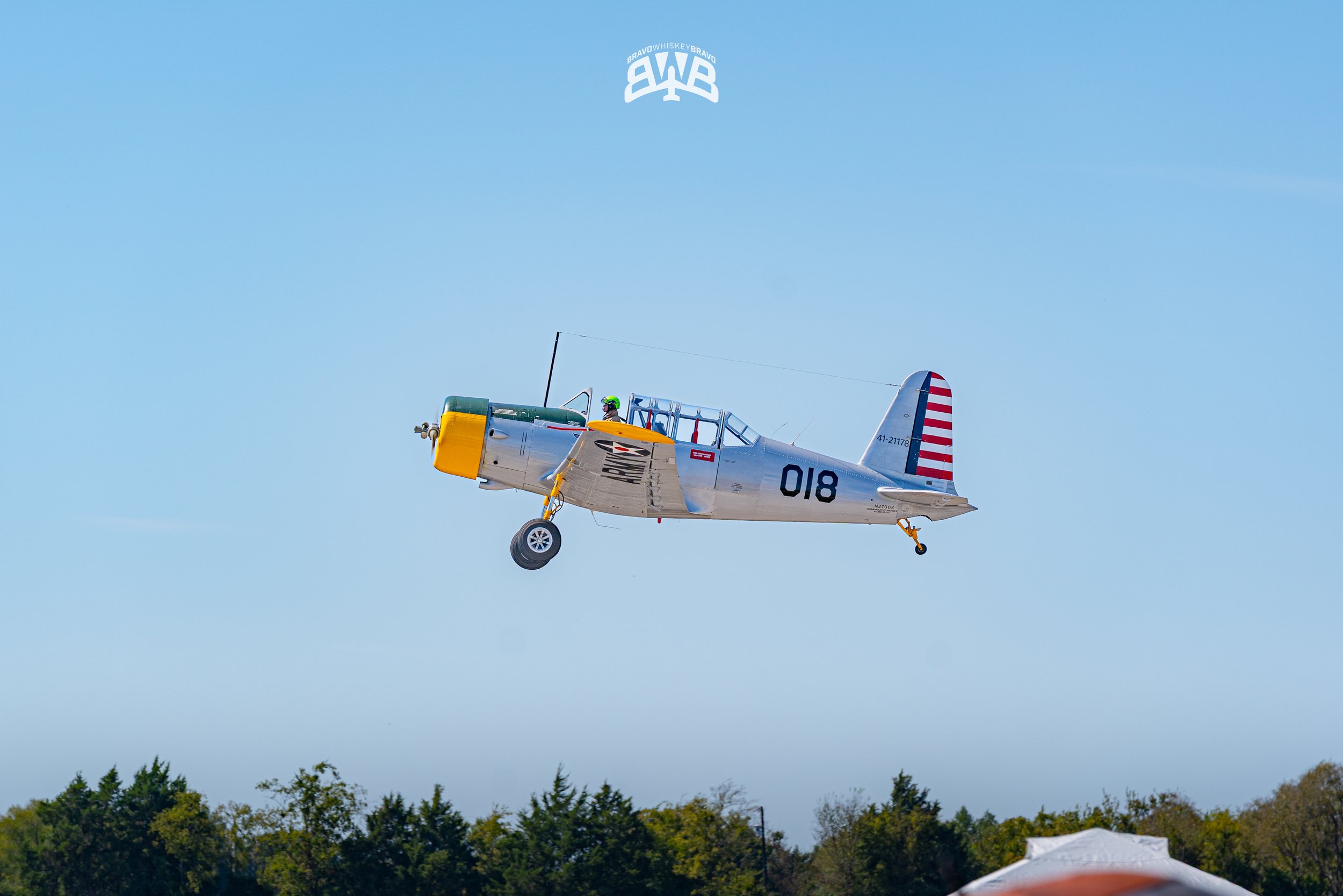 A vintage aircraft flying in the sky above trees, with the American flag design on the tail.