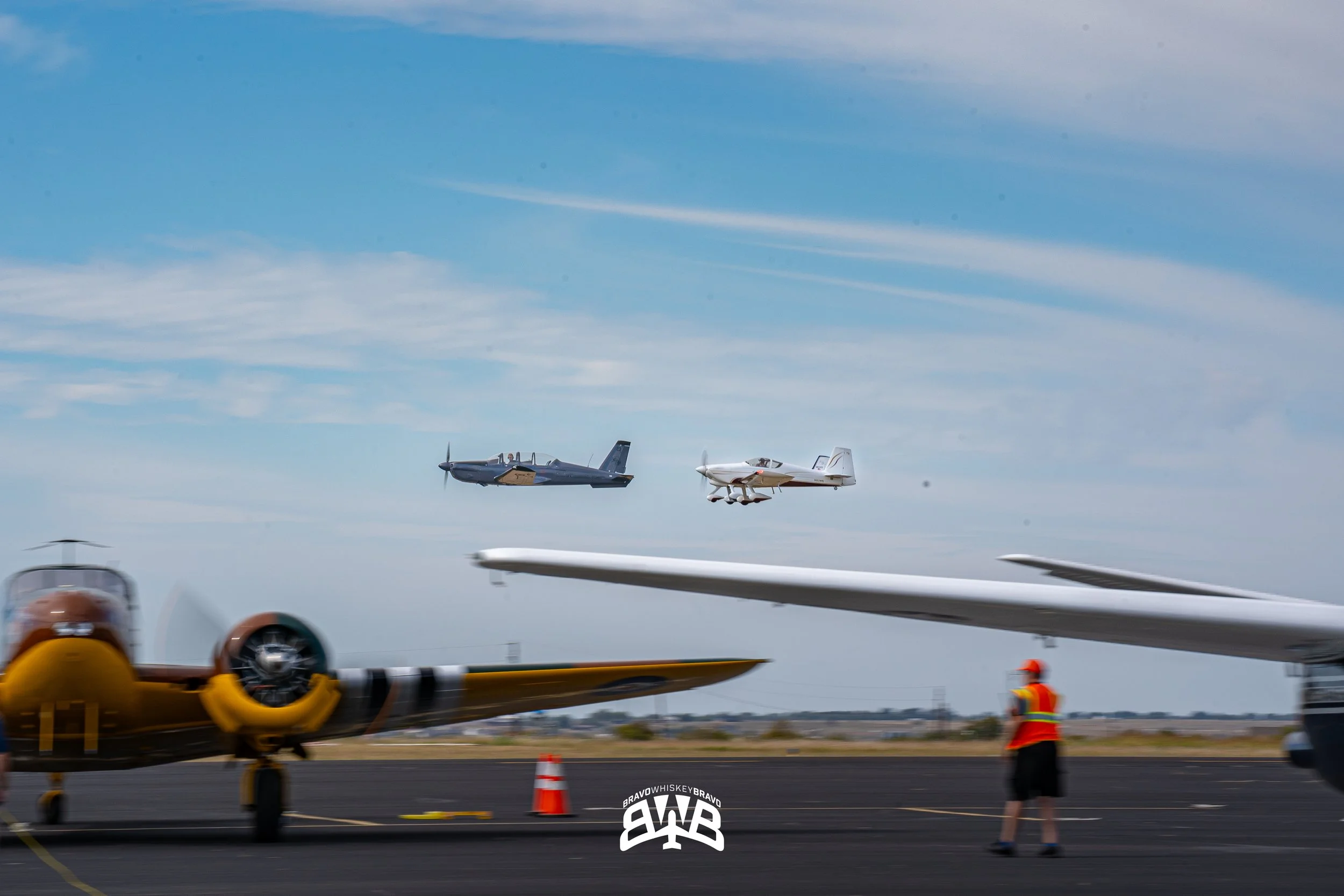 Two vintage aircraft flying in formation above an airfield, with one person in a safety vest and hat observing on the ground. The scene includes a blurred propeller plane in the foreground, and a logo at the bottom center.