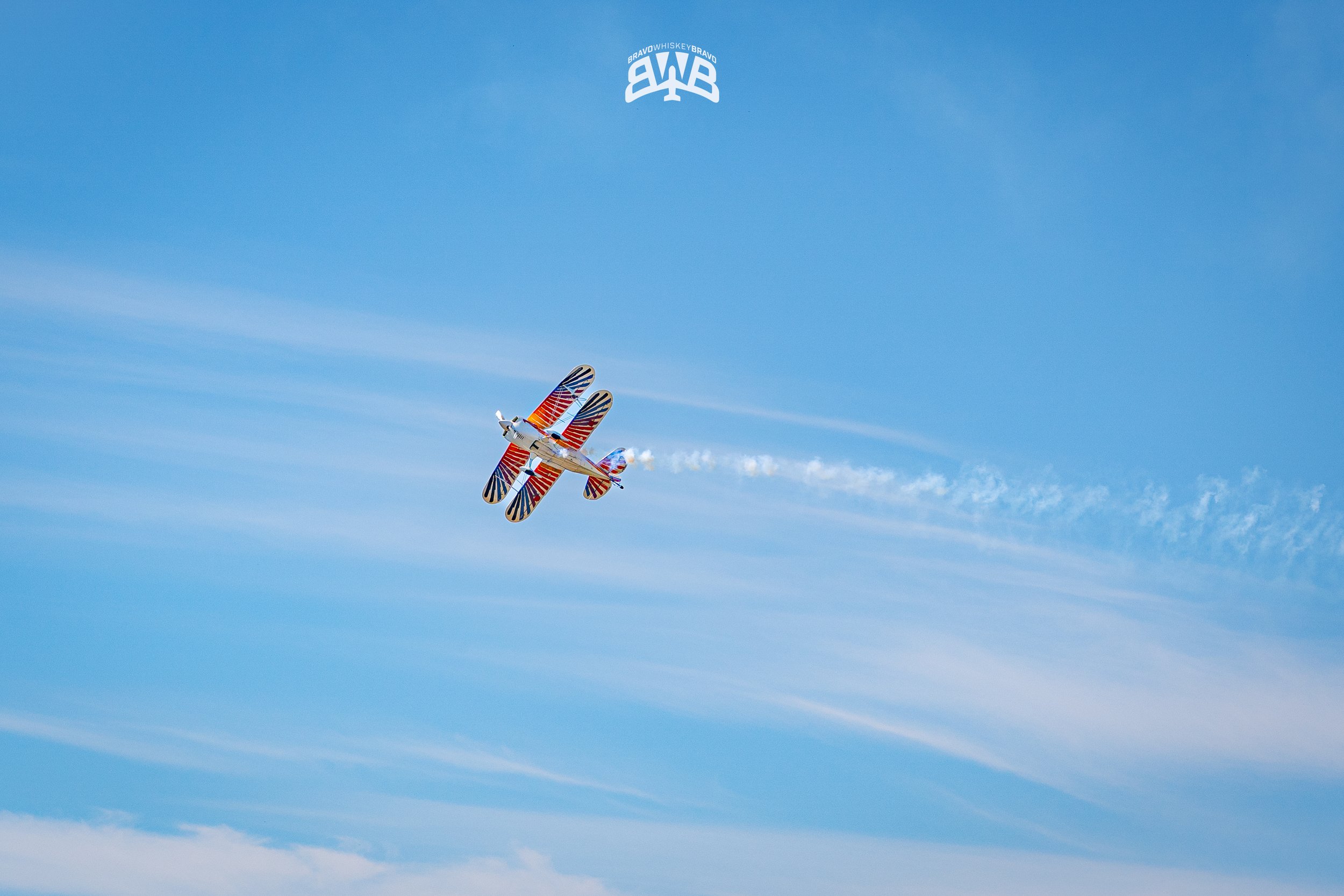 A biplane with red, white, and blue patriotic patterns flying in a clear blue sky, leaving a white smoke trail behind.
