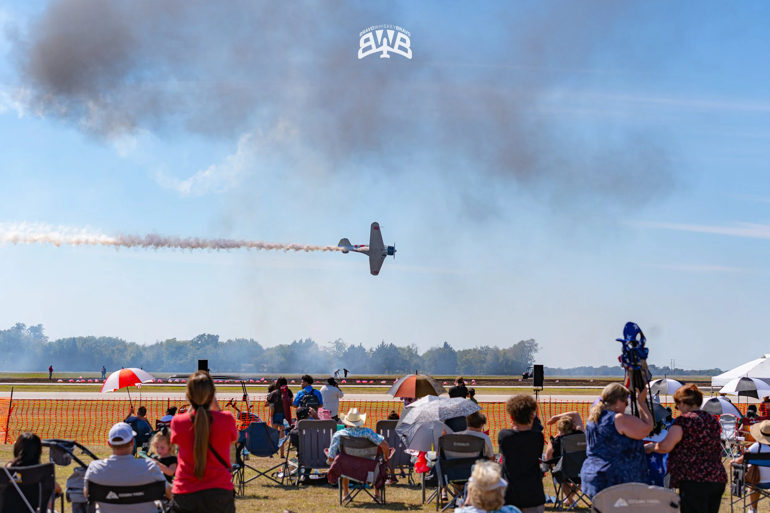 An airshow with a crowd watching a vintage aircraft perform aerobatics, leaving smoke trails in the sky, while some spectators hold umbrellas.