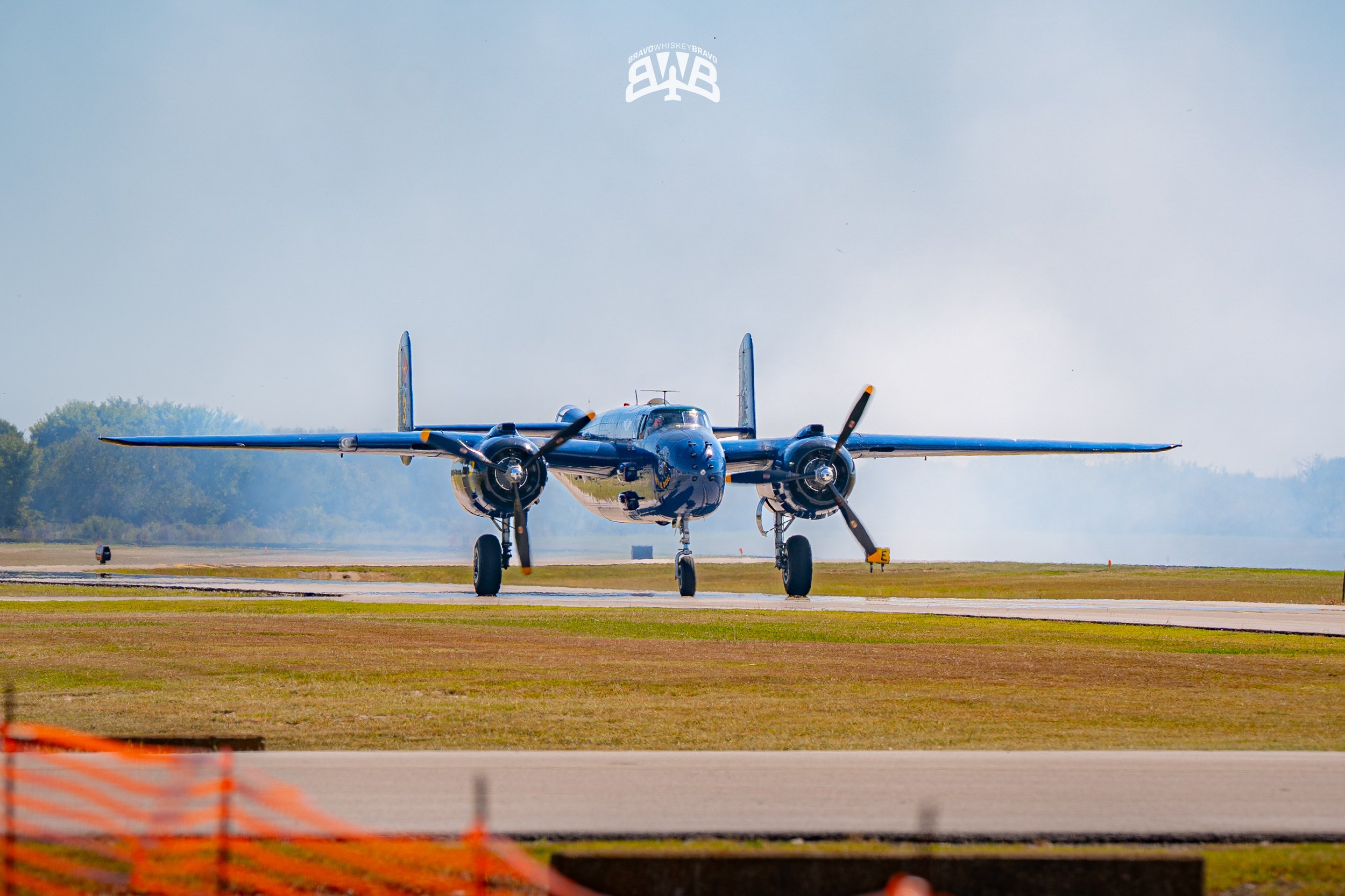 A vintage twin-engine propeller aircraft on a runway with trees in the background.
