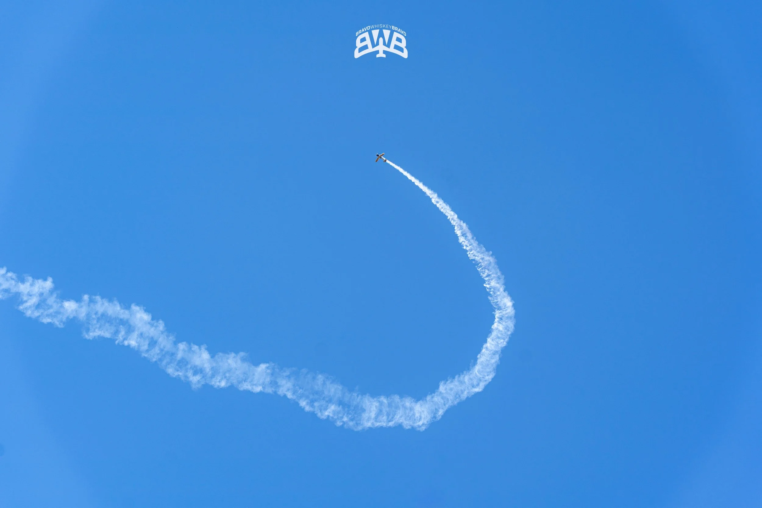 A small airplane performing an aerial stunt, leaving a white contrail in a blue sky with a few clouds, and a logo with the text 'BRAVOWHISKEYBRAVO' at the top.