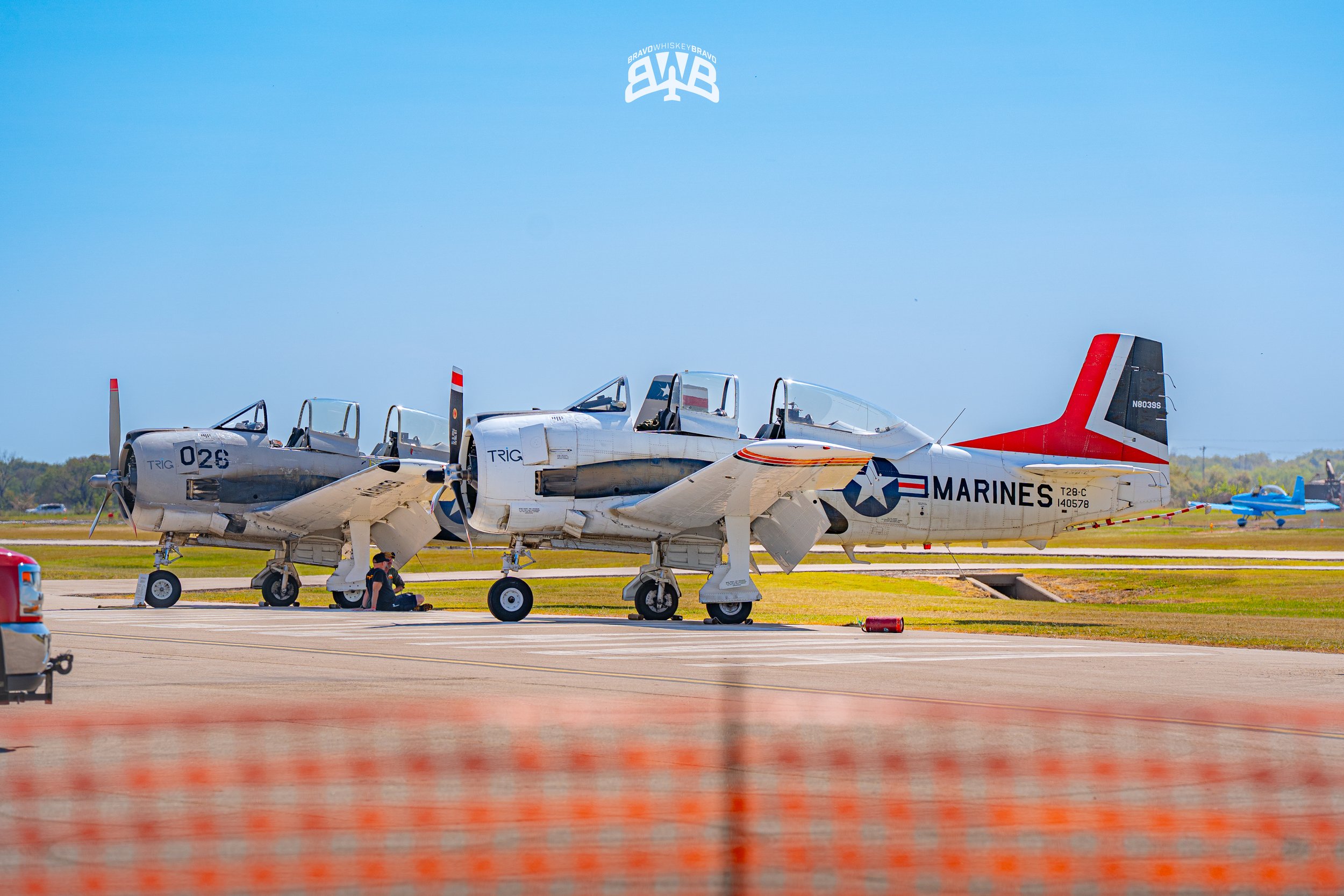 Two vintage US Marine Corps fighter jets parked on tarmac under blue sky, with a person kneeling near the nose of one aircraft.