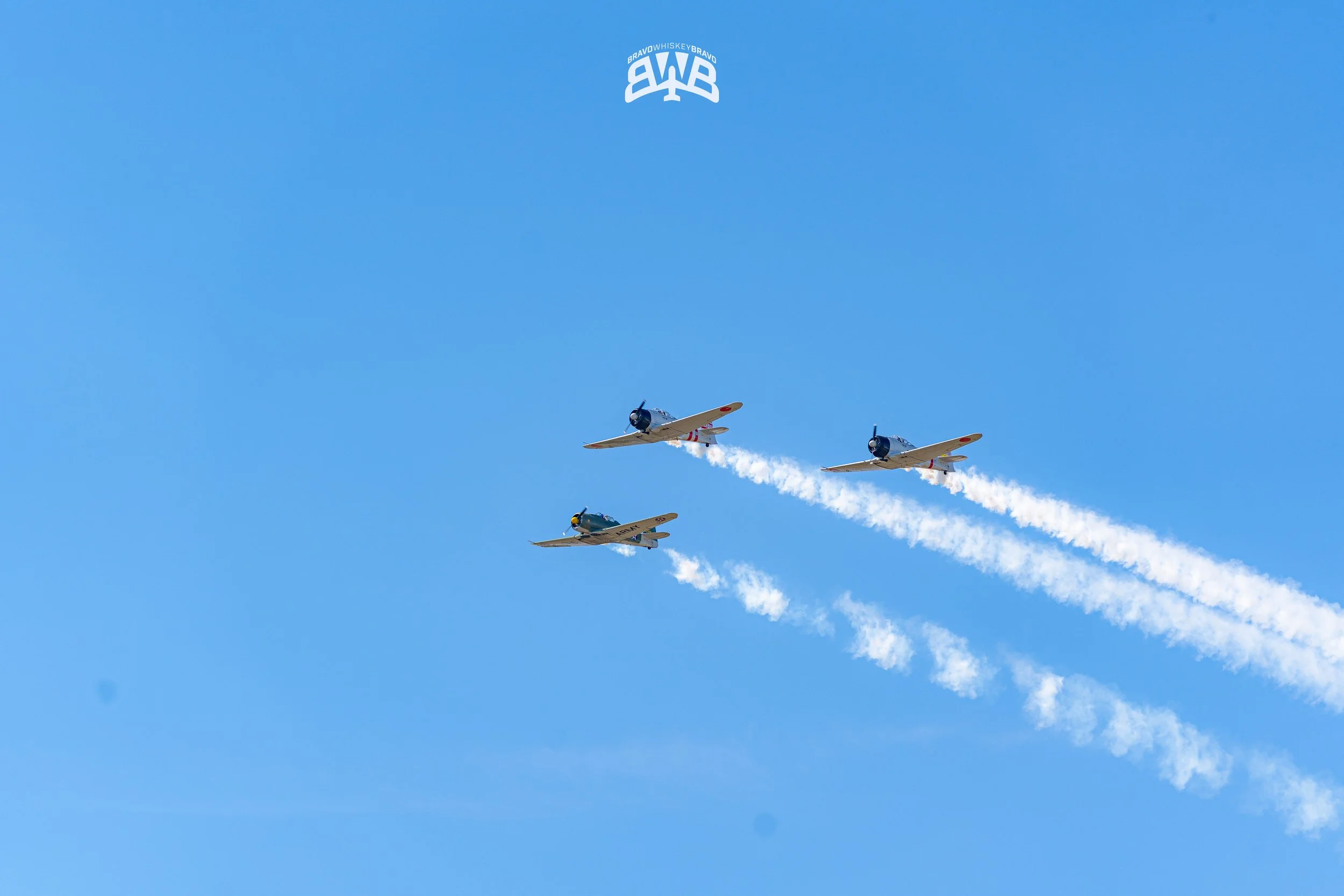 Three vintage fighter planes flying in formation in a clear blue sky, leaving white smoke trails behind them.