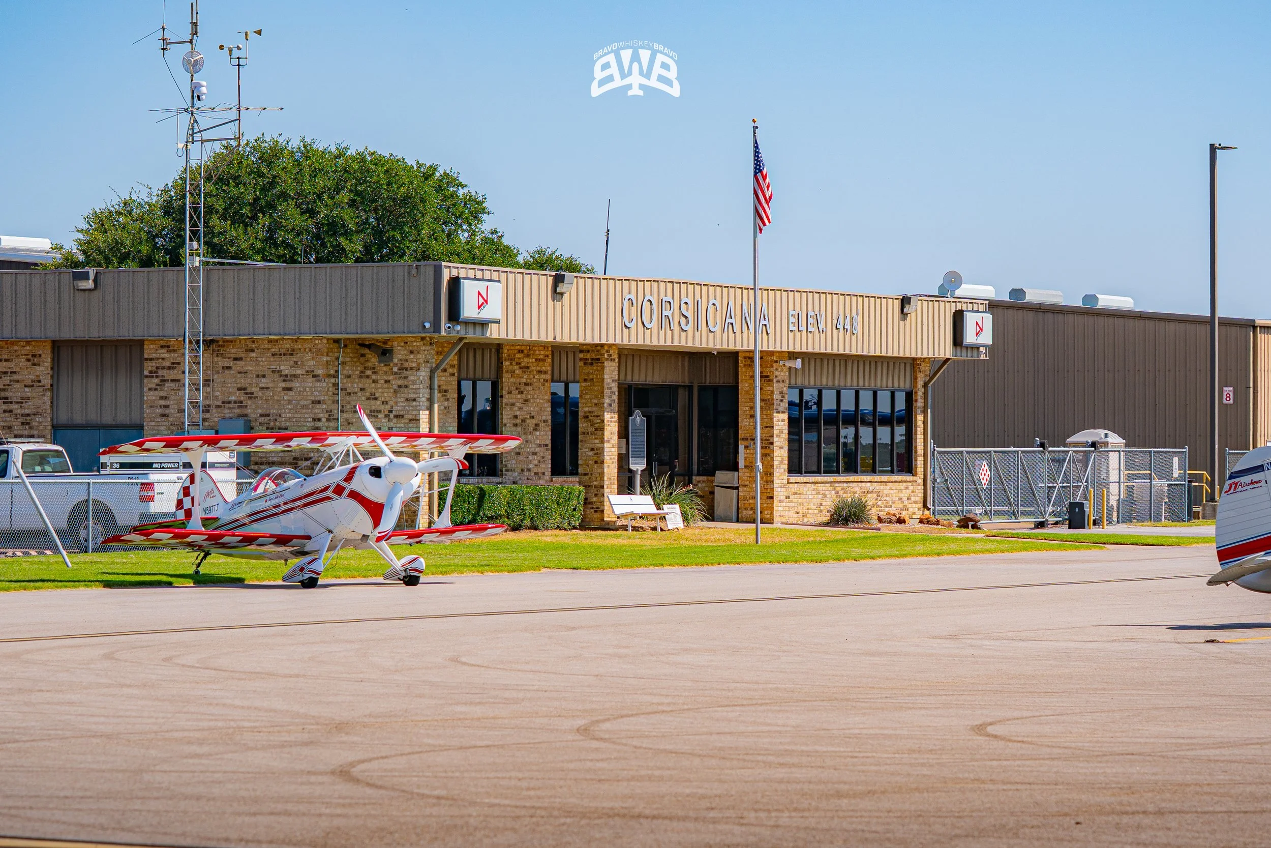 Small aircraft parked on the tarmac outside a beige brick building labeled "CORSICANA ELEV 448" with an American flag, utility poles, and trees in the background.