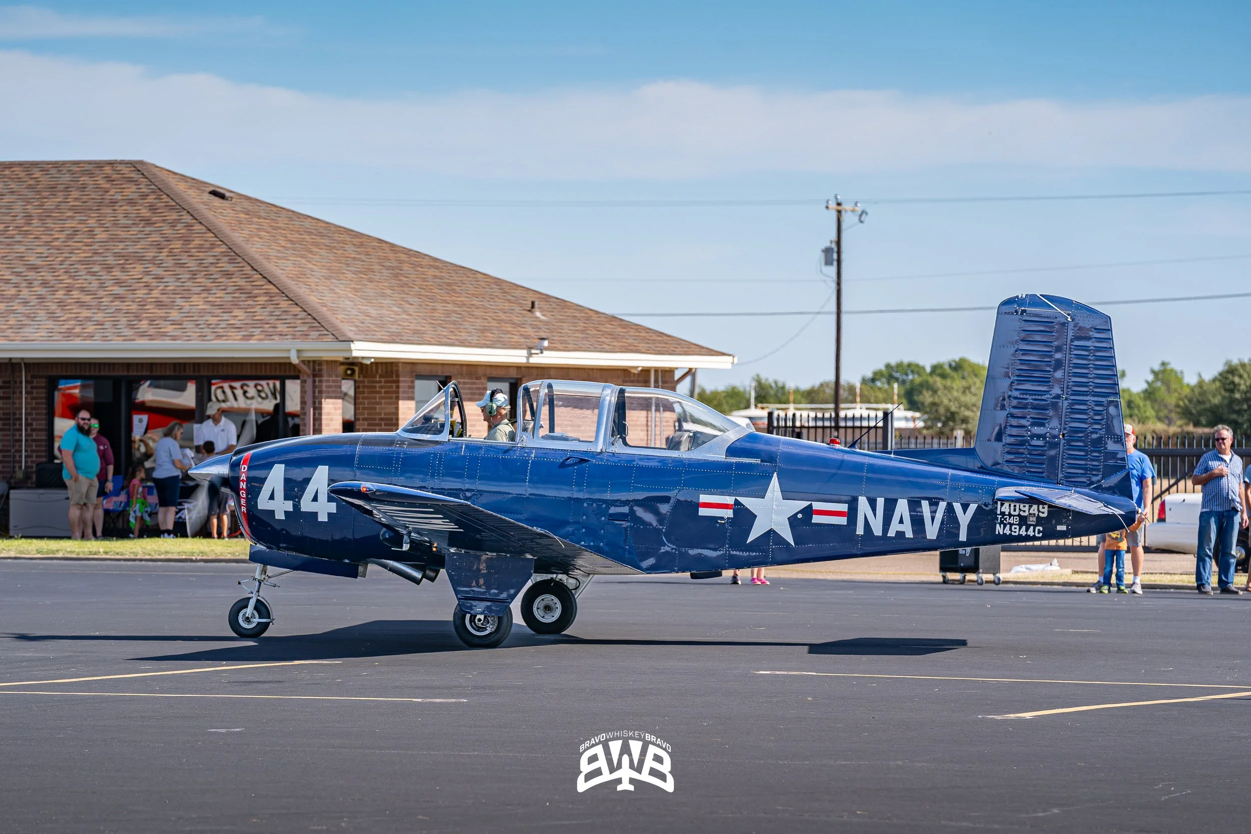 A vintage Navy blue aircraft with the number 44 on its side, parked on an asphalt surface at an event with spectators in the background and a building behind.