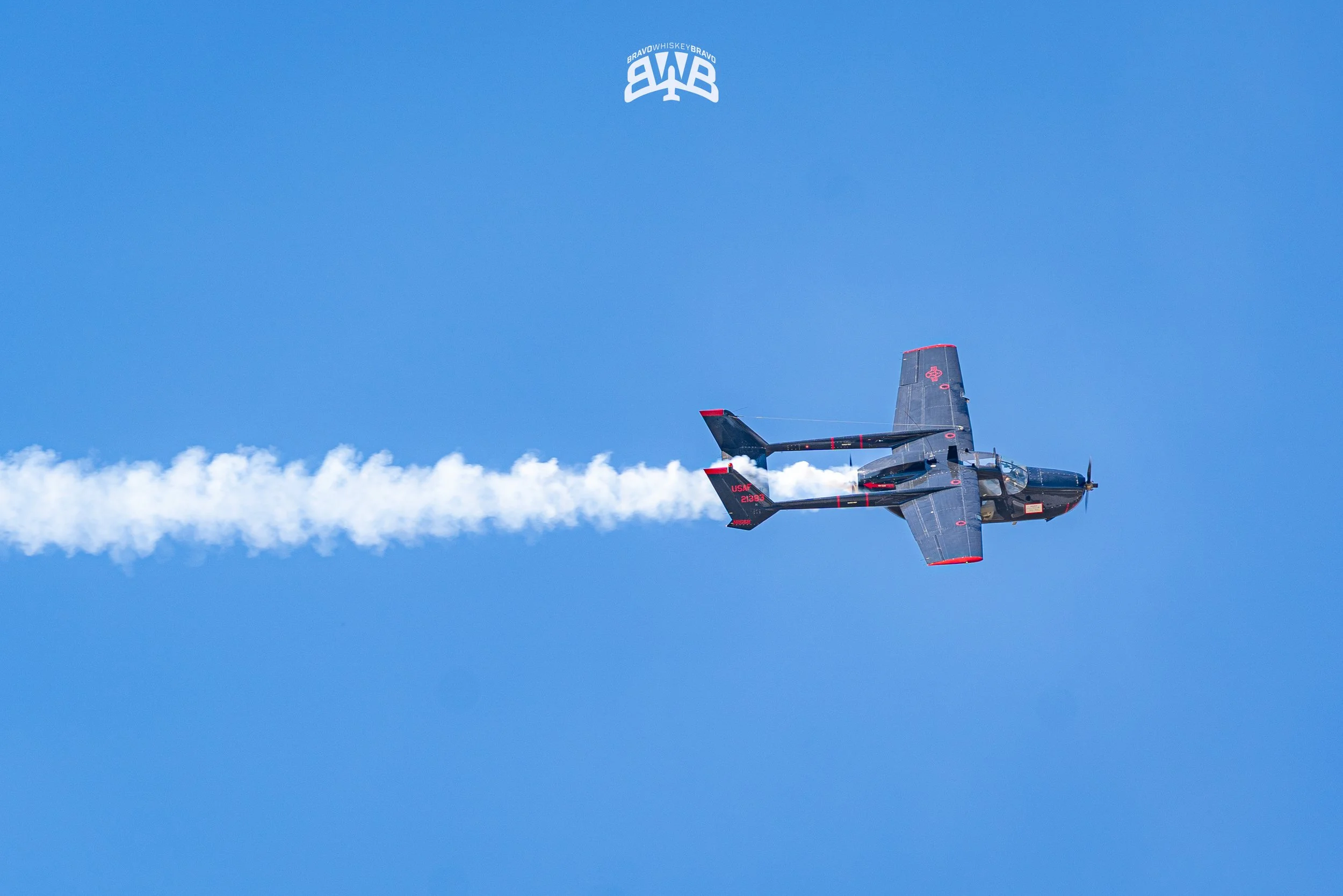 A small jet aircraft flying in clear blue sky, leaving behind white smoke trail, with 'Bravowhiskey' logo upside down at the top of the image.