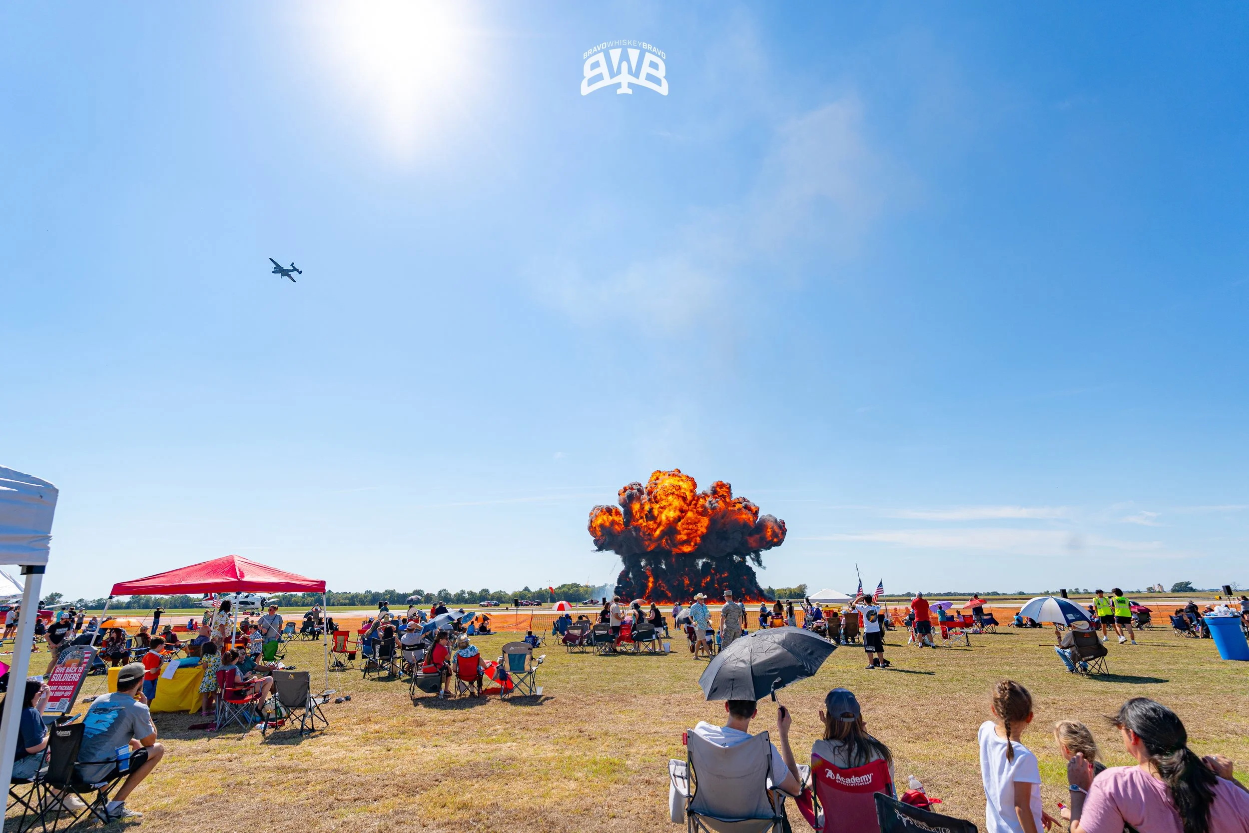 People seated on chairs and on the ground at an outdoor event, watching an airplane crash into a large explosion in the sky during the daytime.