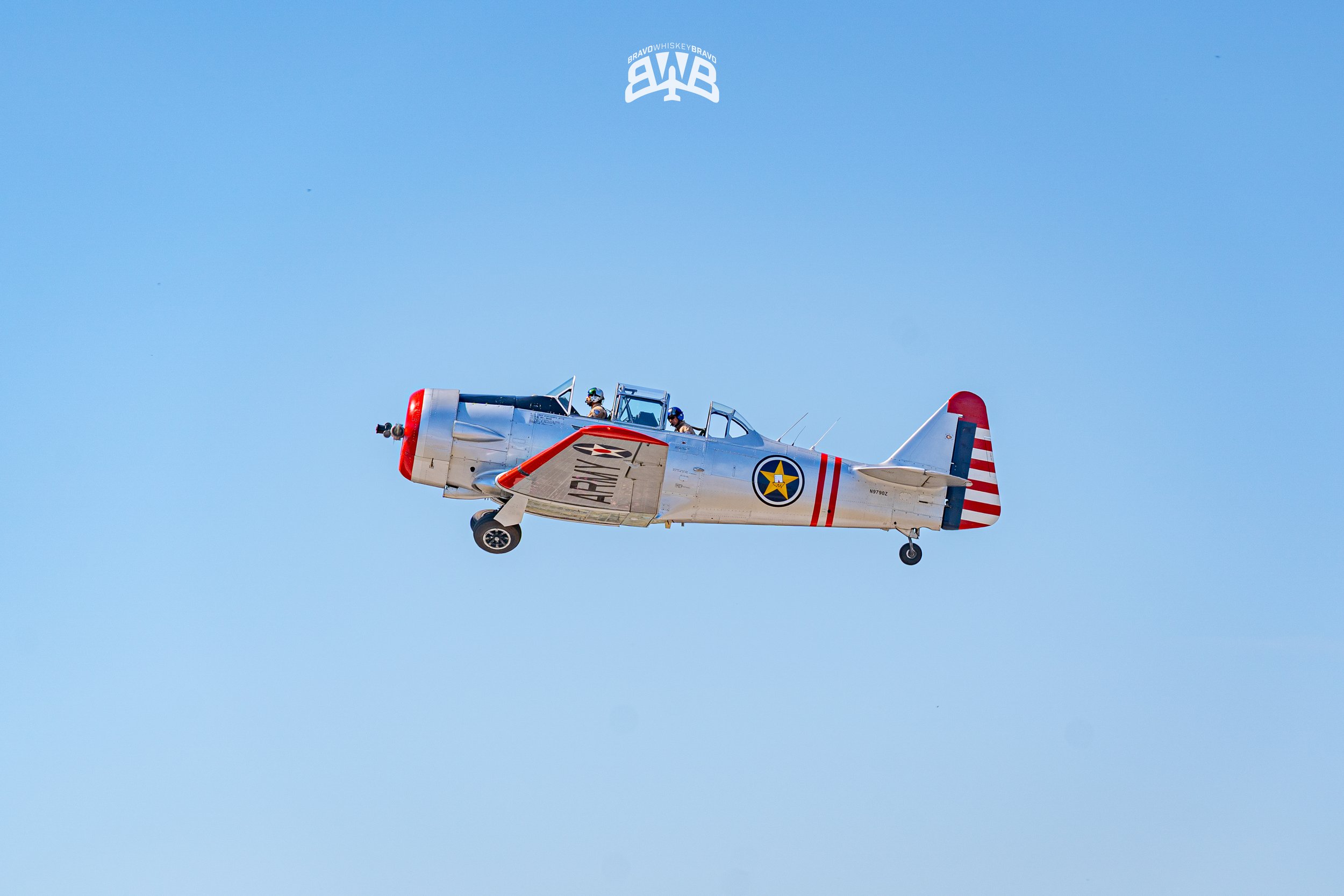 A silver vintage airplane with red and white striped tail flying against a clear blue sky.