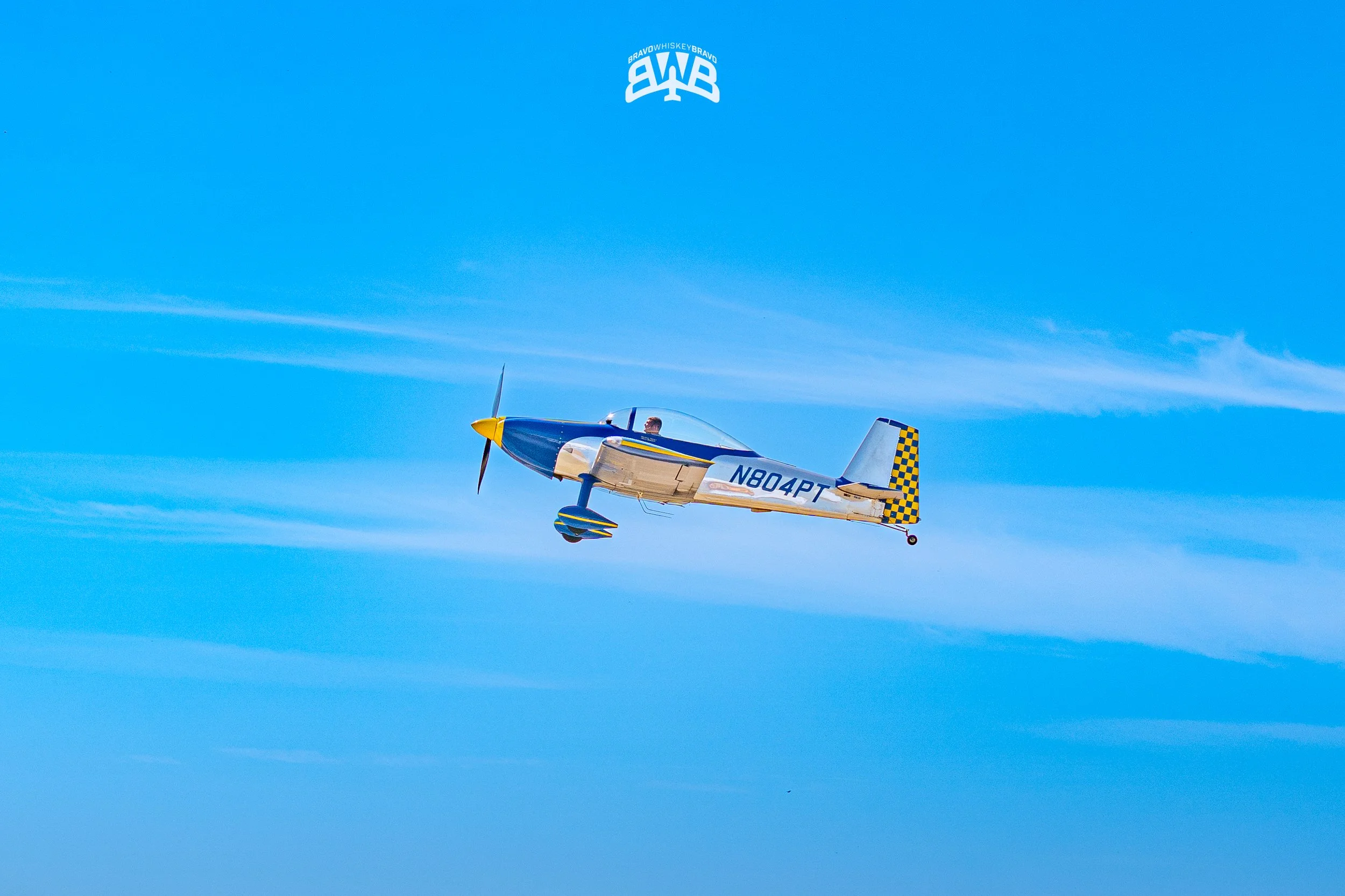 A small propeller airplane with a blue, yellow, and white color scheme flying through a clear blue sky. The tail has a checkered pattern and the registration N804PT is visible.