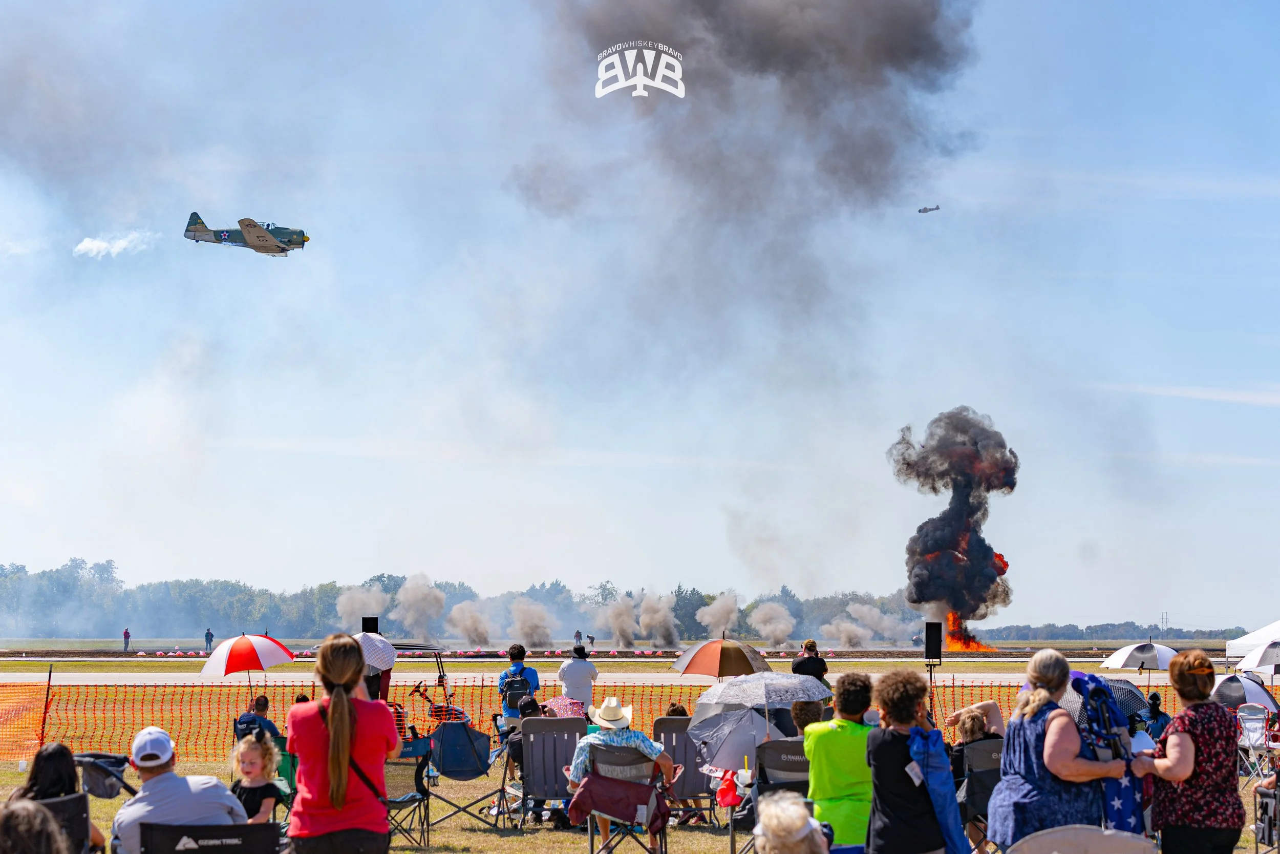 Aerial view of an aircraft performing a smoke display at an air show, with smoke and fire explosions on the ground, and spectators watching from the foreground under umbrellas.