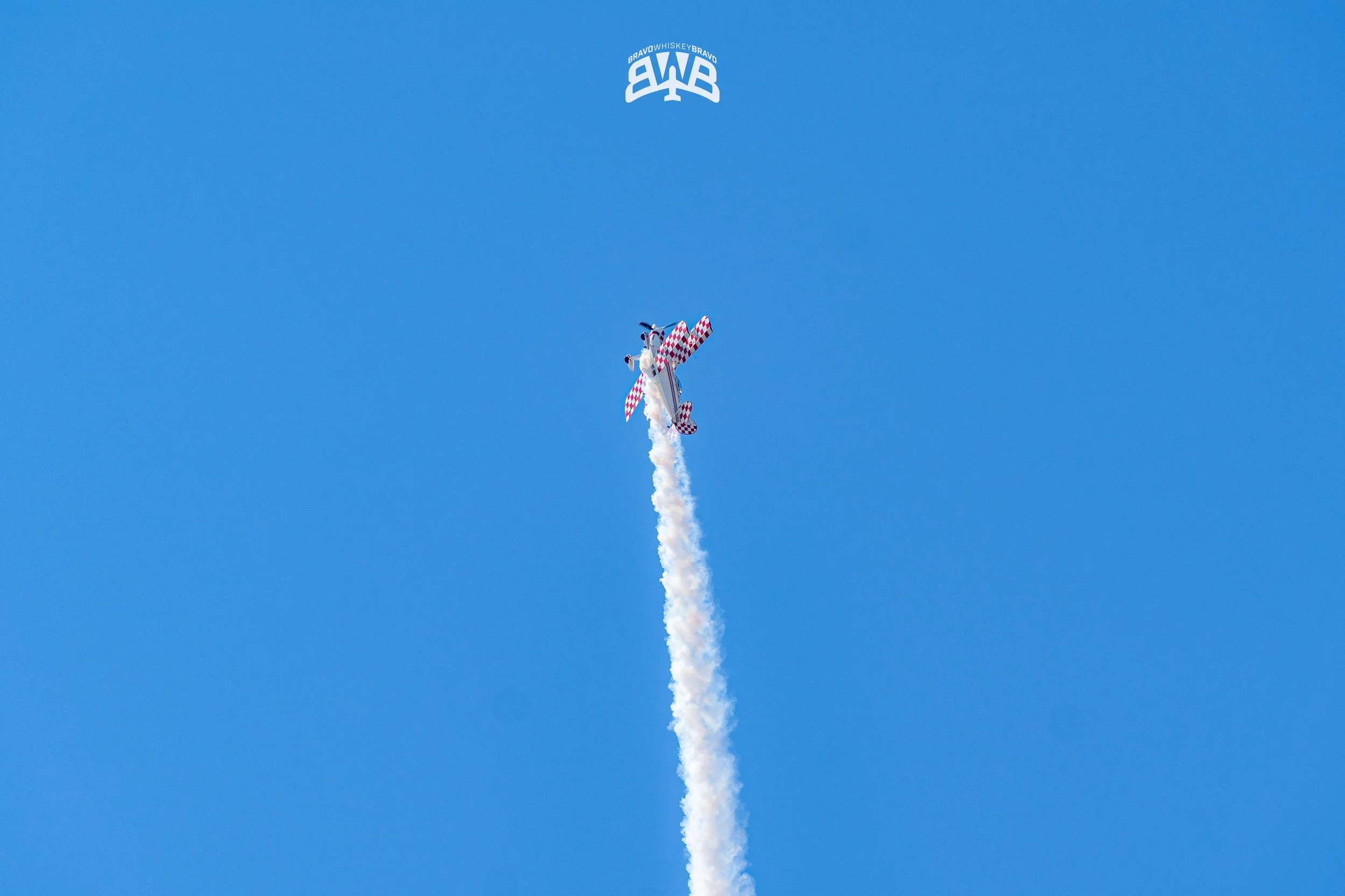 An airplane performing a stunt in the sky with a trail of white smoke behind it, and a logo with the letters 'BRAVO WHISKEY BRAVO' at the top of the image.