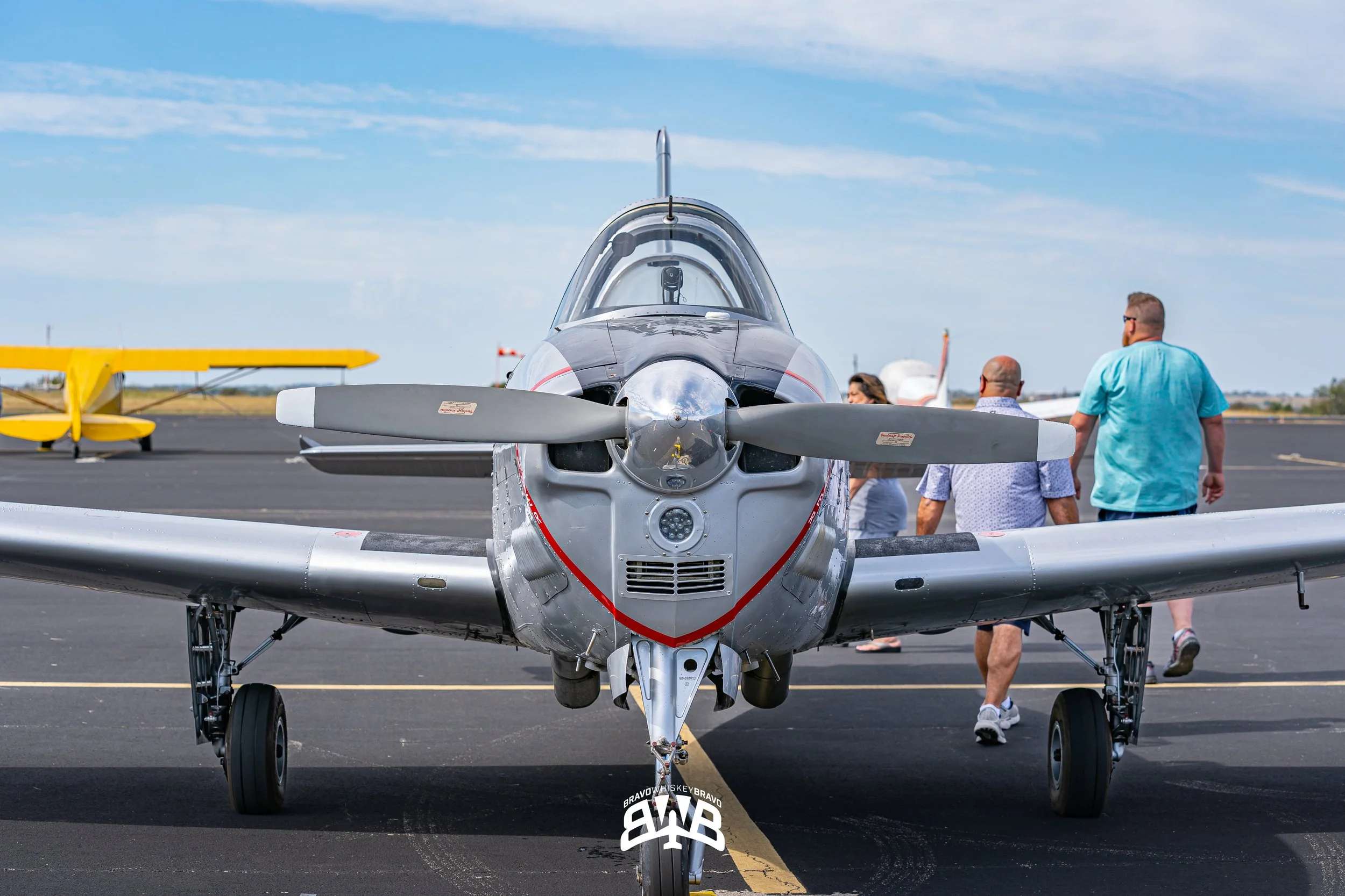 Front view of a silver fighter jet parked on an airstrip with several people walking behind it and a yellow plane in the background under a blue sky with clouds.