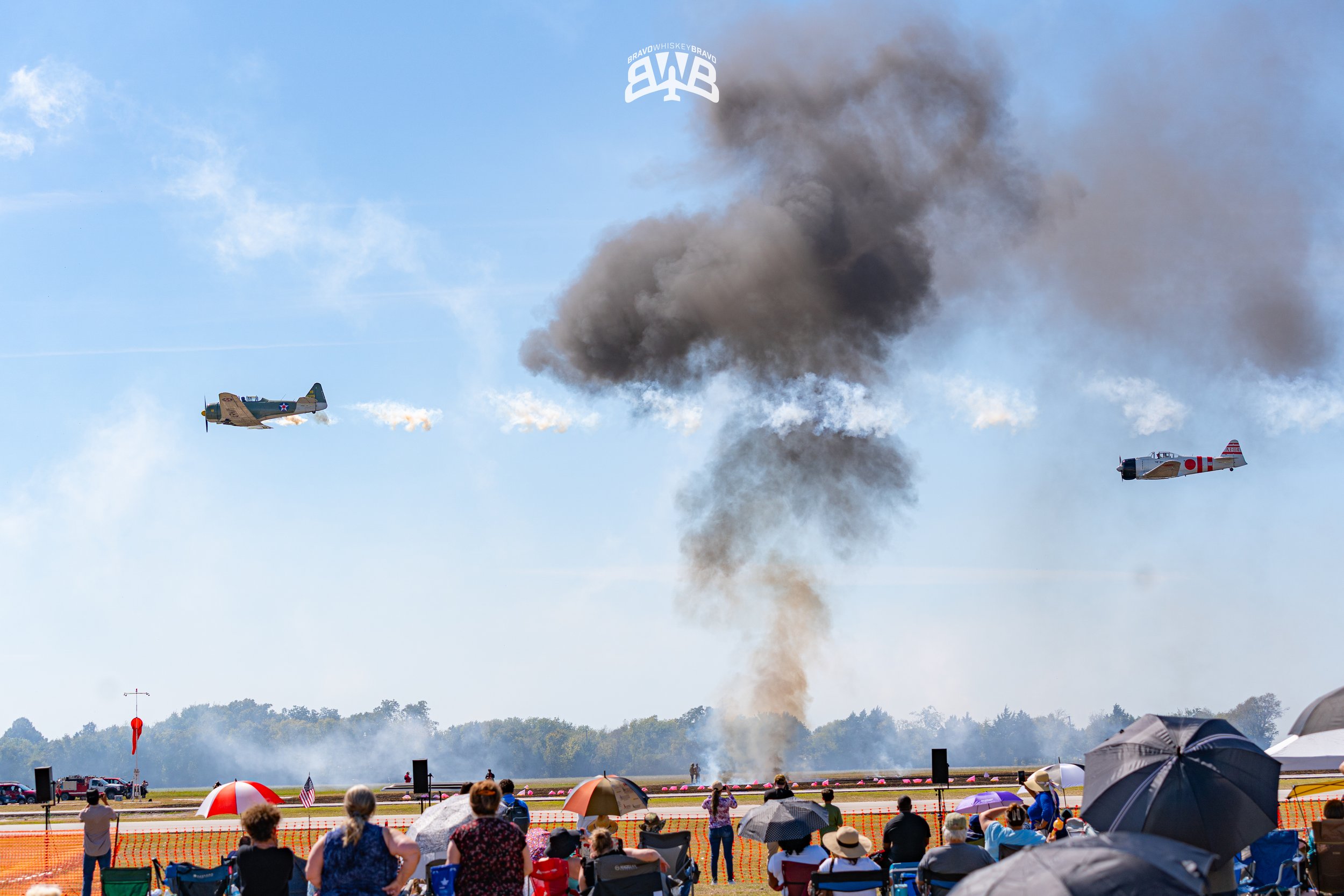 Two vintage planes flying in the sky with smoke trails and an explosion occurring in midair during an air show, with a crowd of spectators watching on the ground.