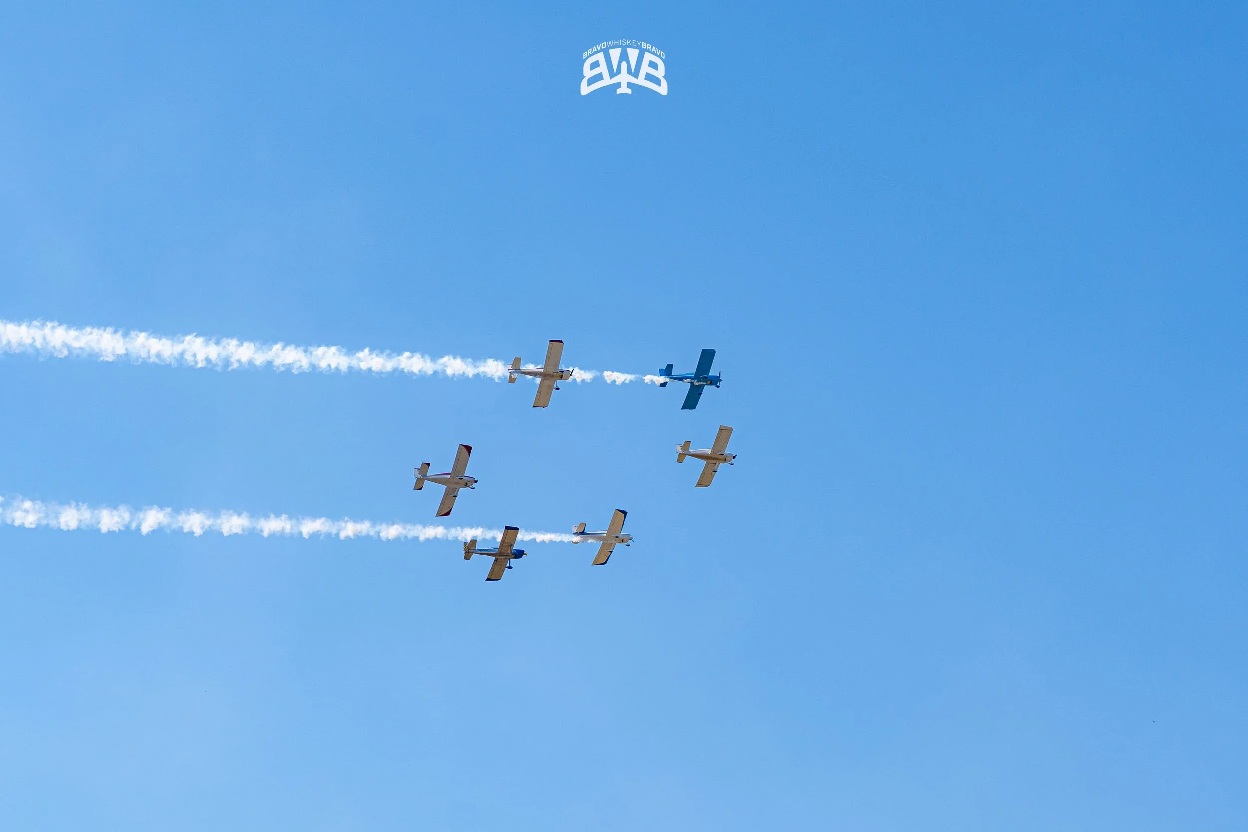 Six airplanes flying in formation against a clear blue sky, leaving white smoke trails behind.