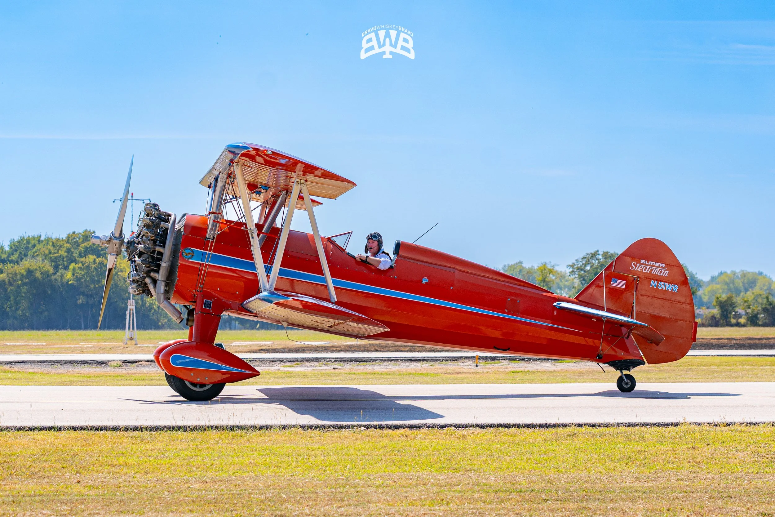 A vintage red biplane with blue and white stripes on the fuselage, taxiing on a runway with a pilot in the cockpit wearing goggles and a helmet.
