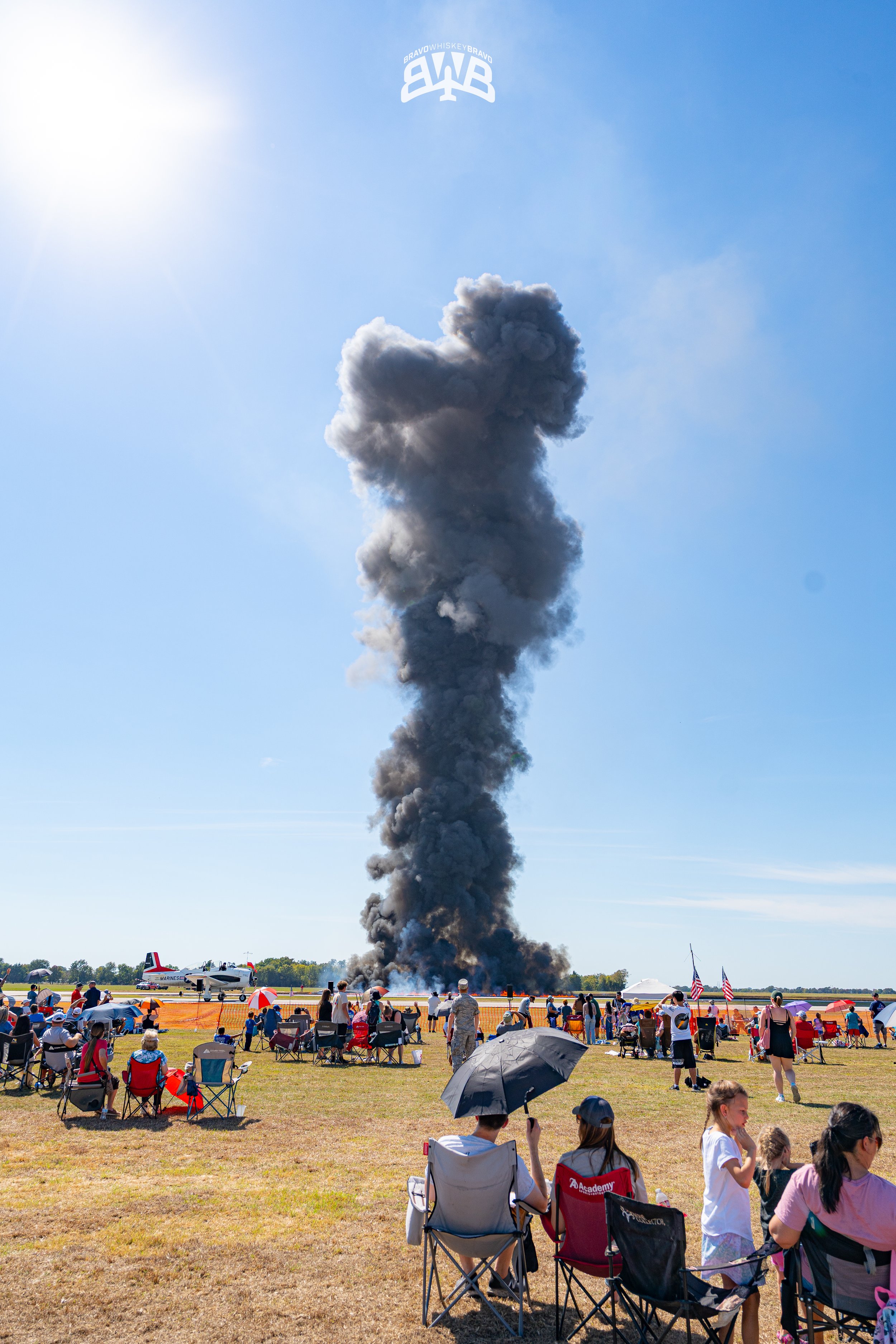 People watching a plane on fire and releasing black smoke at an airshow, with spectators seated on the grass under umbrellas and flags, on a bright sunny day.