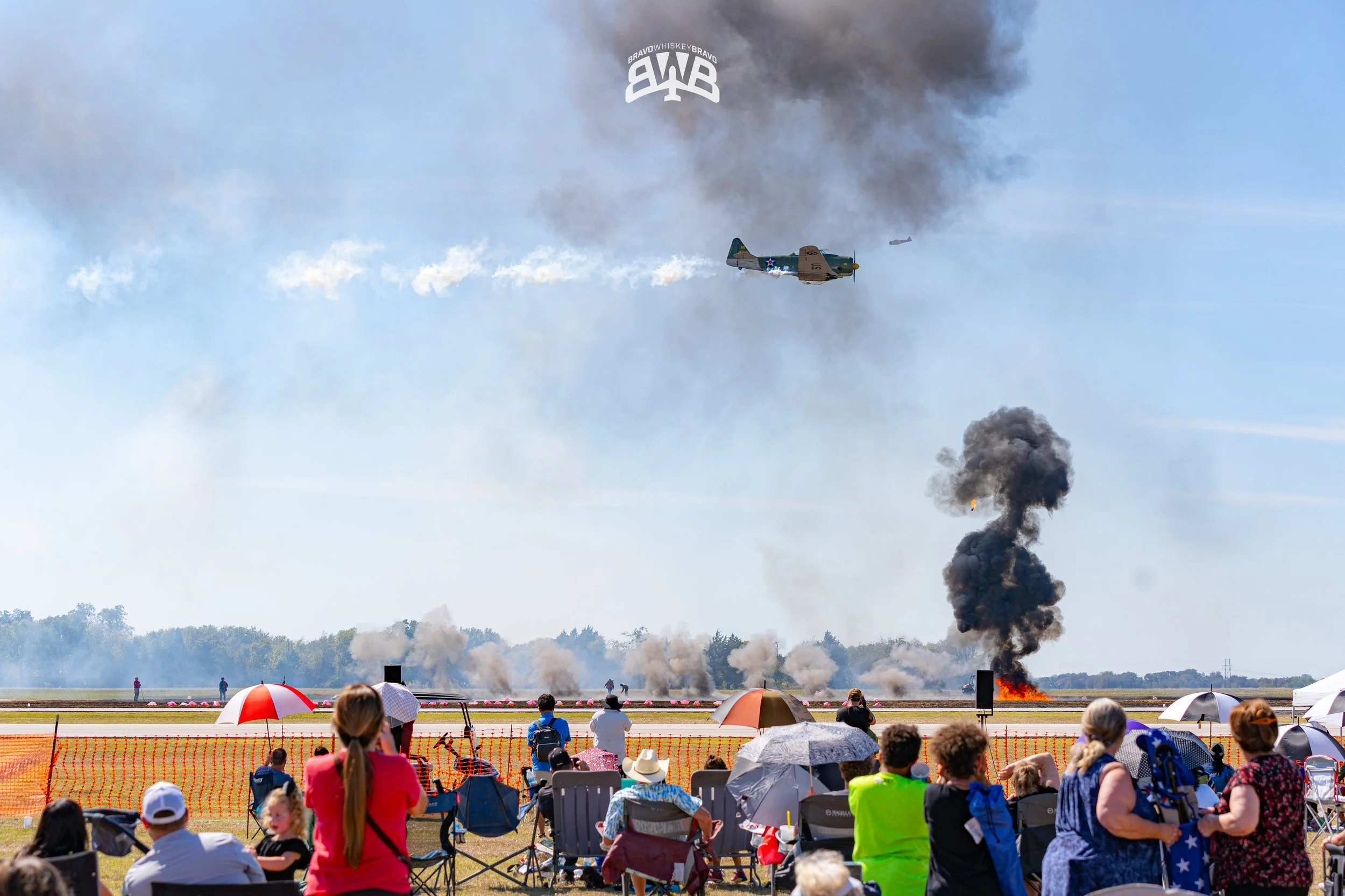 Airplane flying over a crowd at an air show with smoke and fire on the ground.