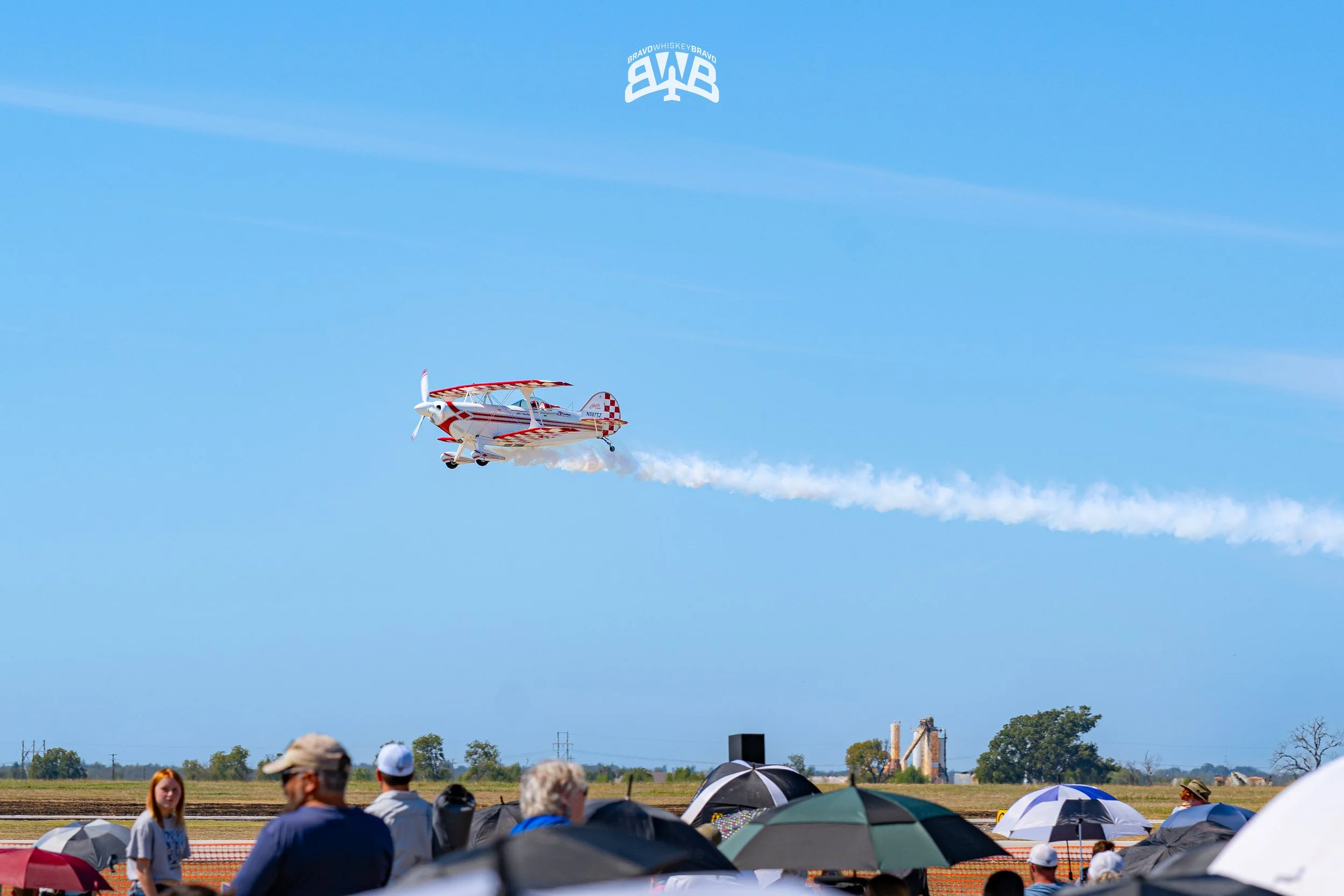 A small airplane flying in the sky, leaving a trail behind, during an airshow with spectators and umbrellas on the ground.