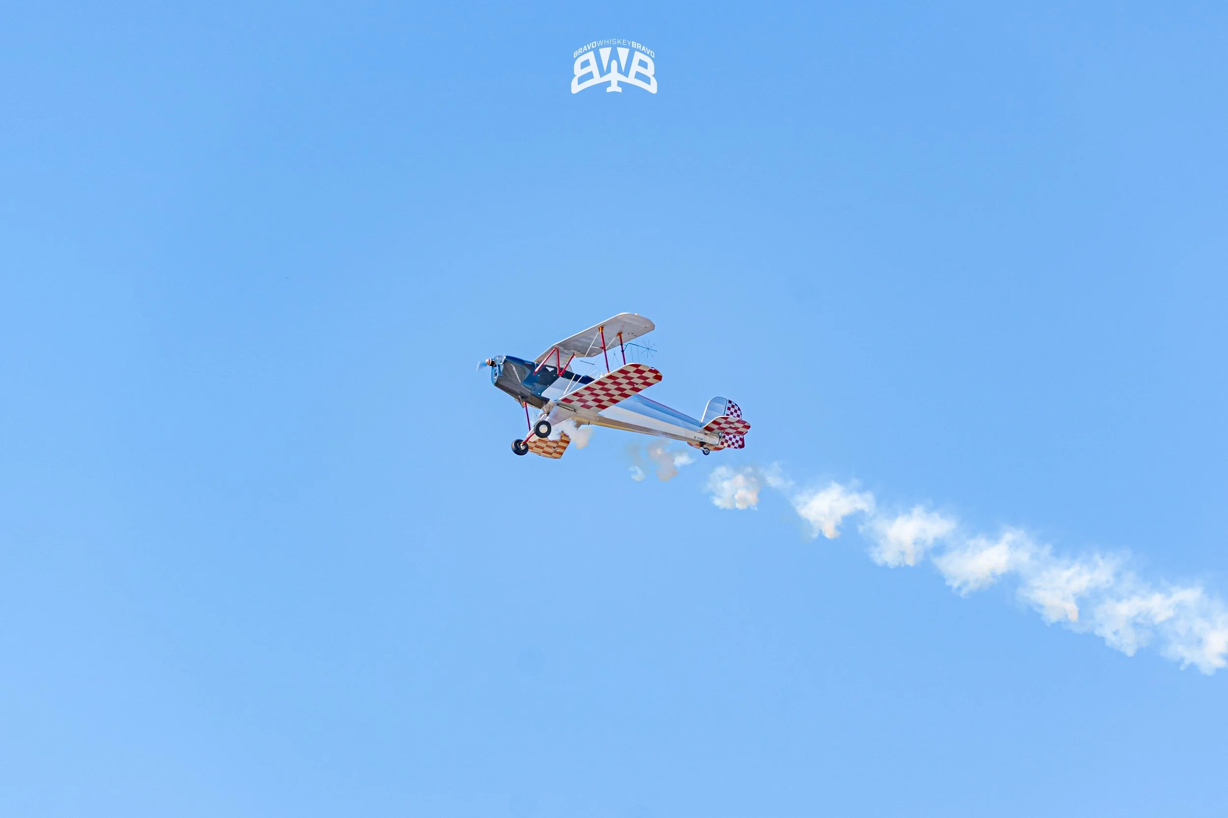 An aerobatic biplane airplane flying in a clear blue sky, leaving white smoke trail behind.