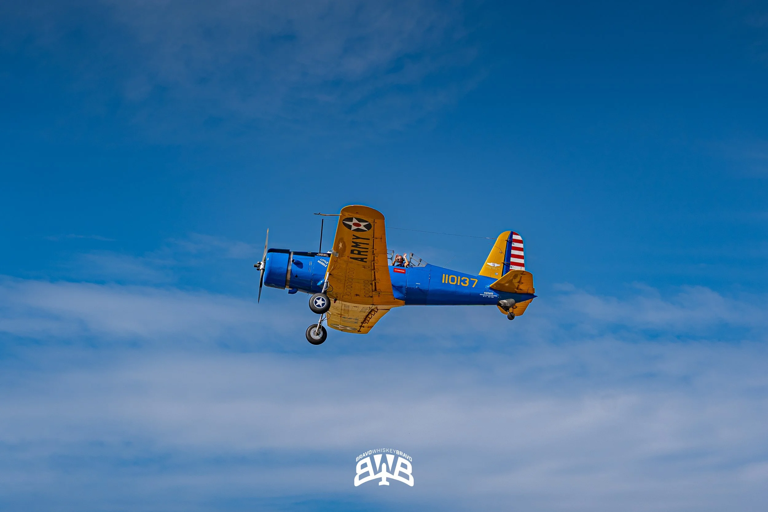A vintage blue and yellow aircraft with army markings flying in a blue sky.