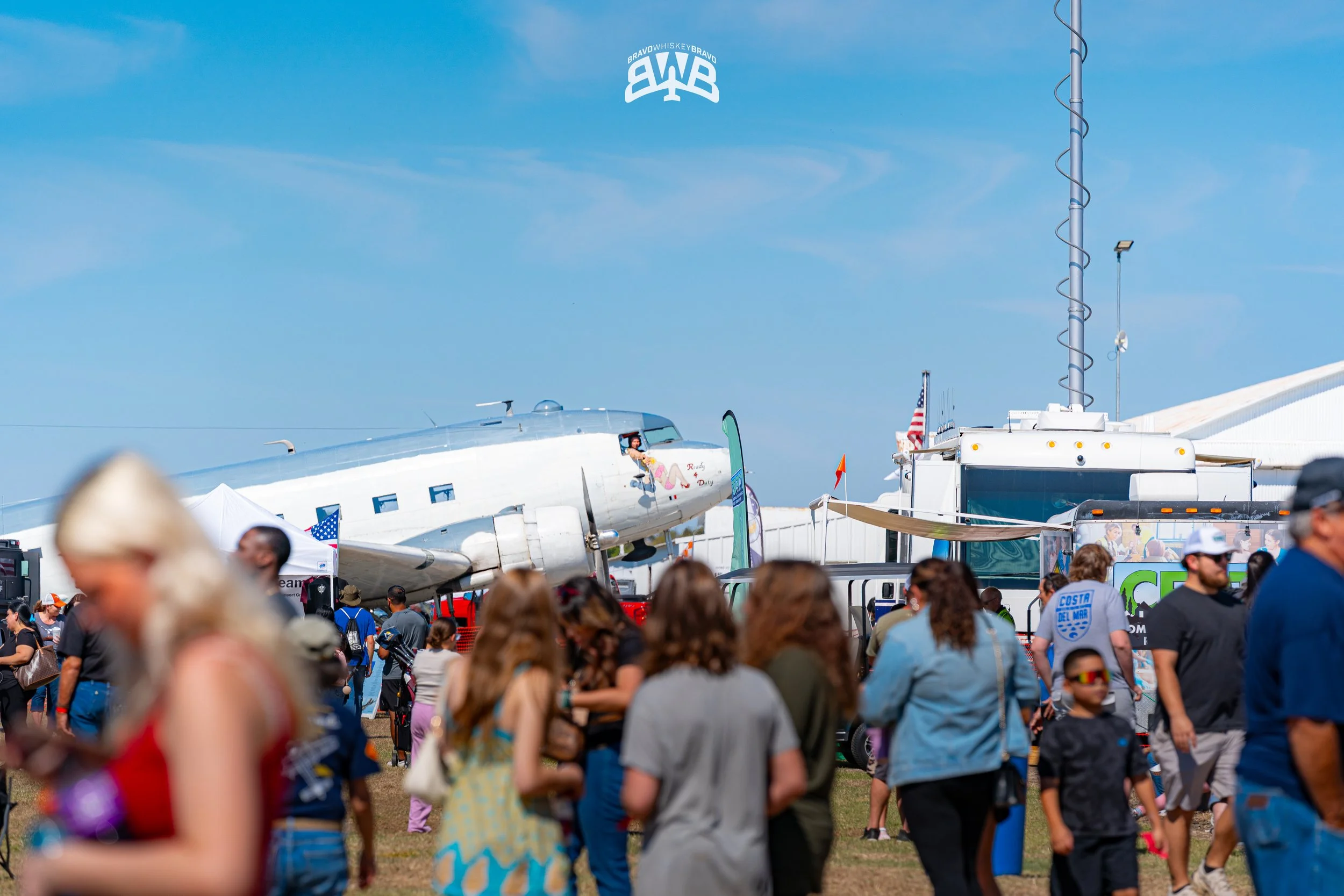 At an outdoor airshow, a crowd of people gathers in front of a vintage aircraft on display, with various tents and flags in the background, and a person visible inside the aircraft's cockpit.