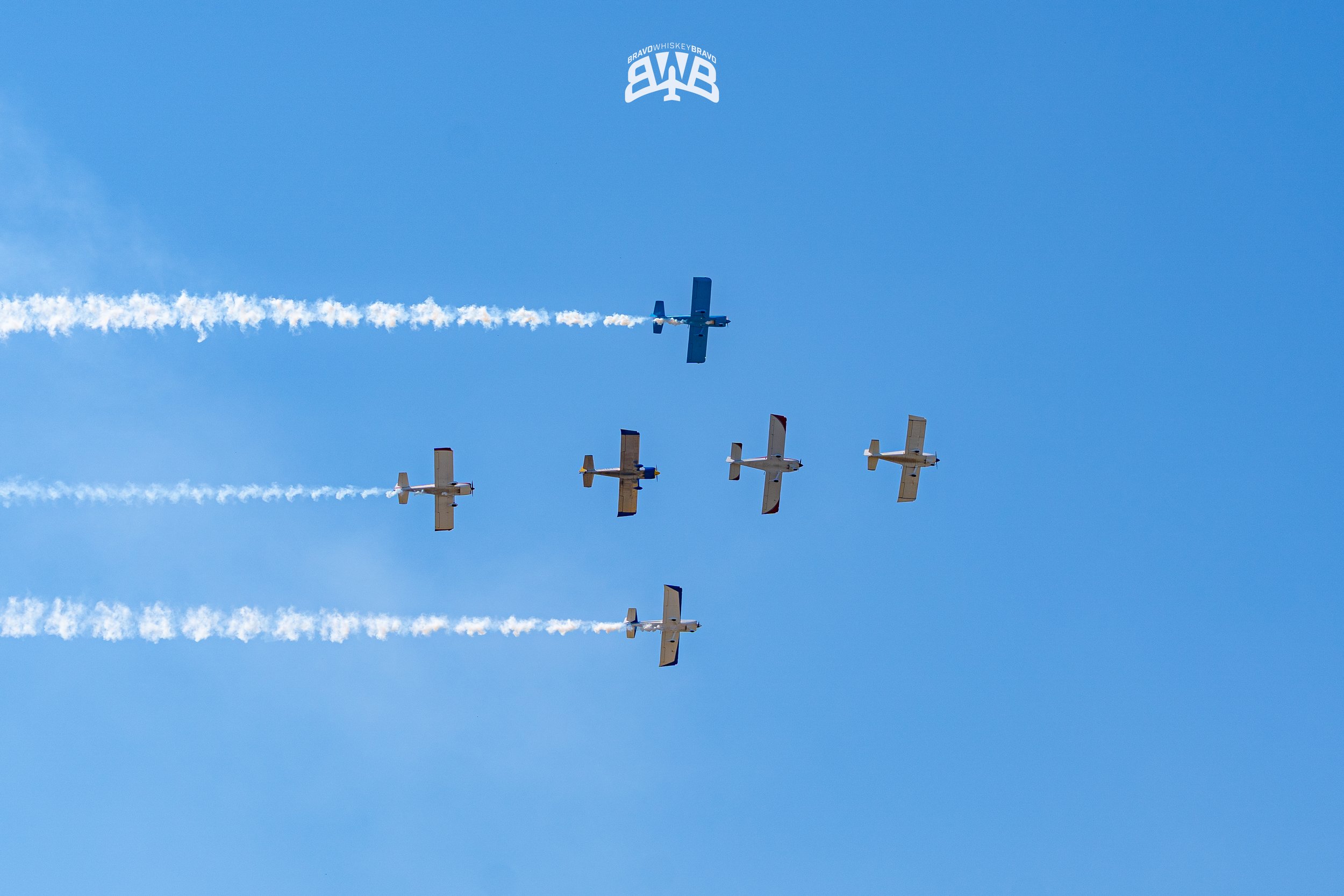 Six airplanes flying in formation against a blue sky, emitting white smoke trails, with one aircraft at the top and five planes below, all aligned in a coordinated air show display.