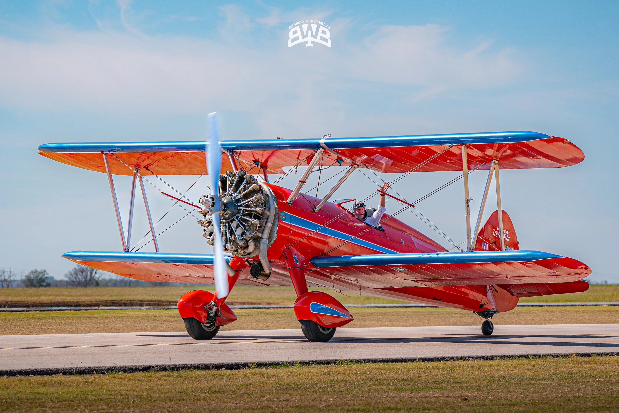 A red biplane with blue accents on a runway, with a person waving from the cockpit.