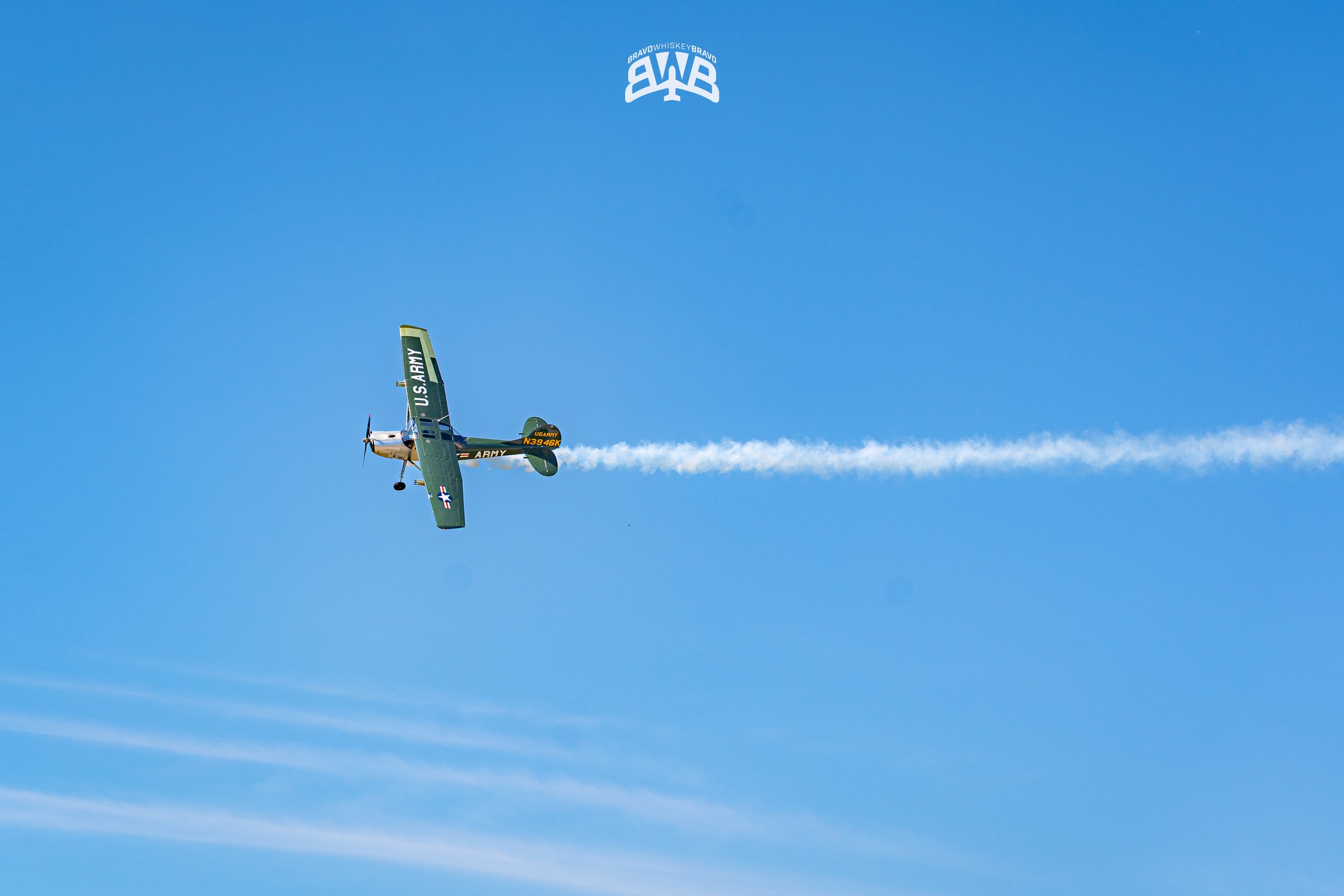 A vintage U.S. Army aircraft flying in a blue sky leaving a white smoke trail behind it.