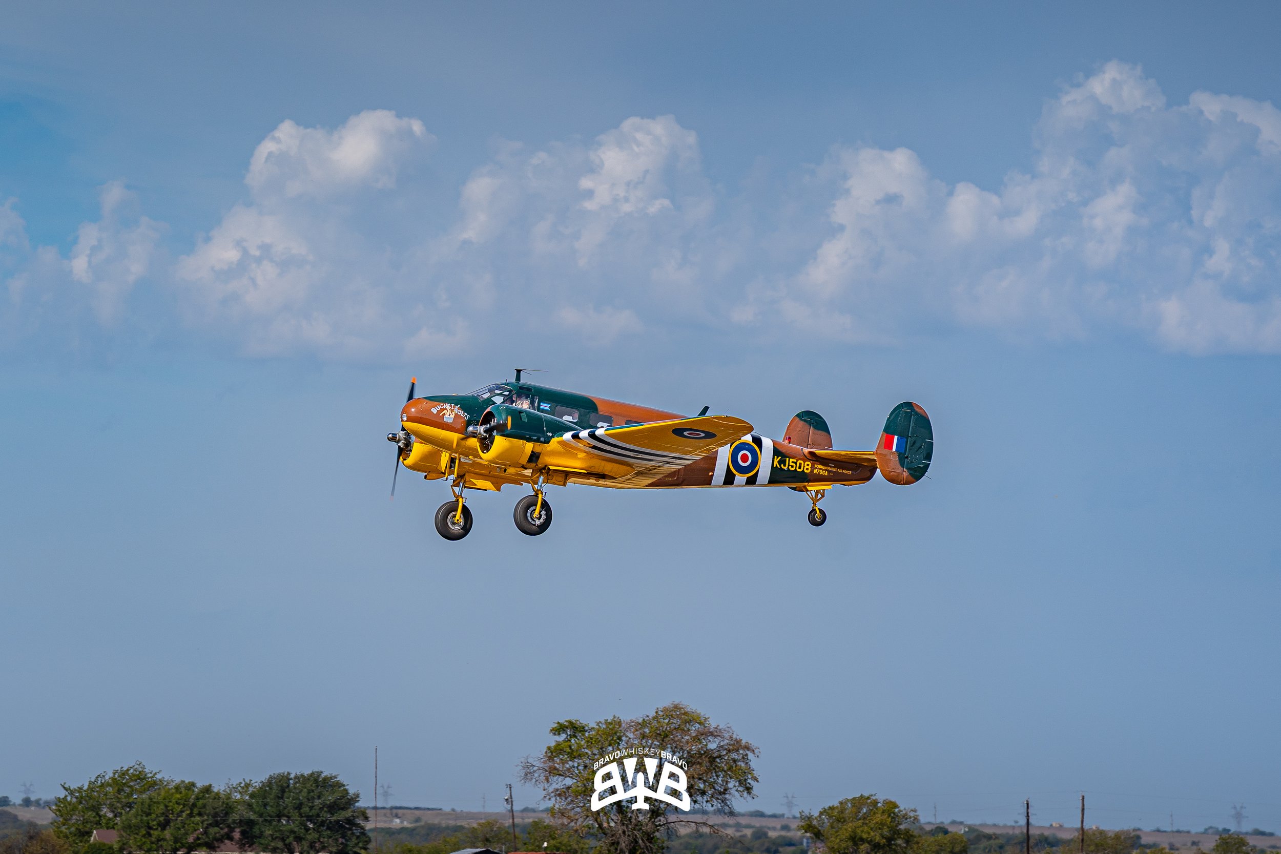 A colorful vintage aircraft flying in the sky above trees and landscape, with a partly cloudy blue sky background.