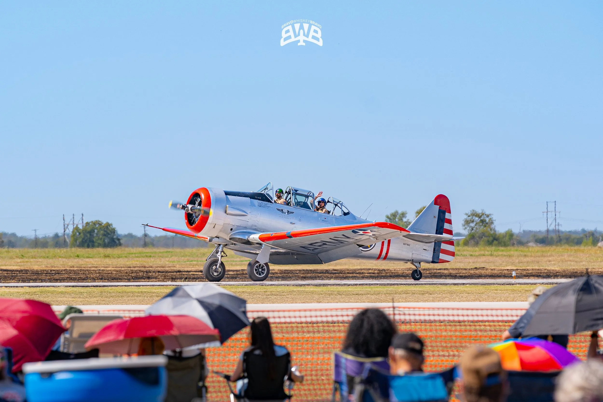 A vintage silver airplane with red accents on the runway at an airshow, with spectators holding umbrellas in the foreground under a clear blue sky.