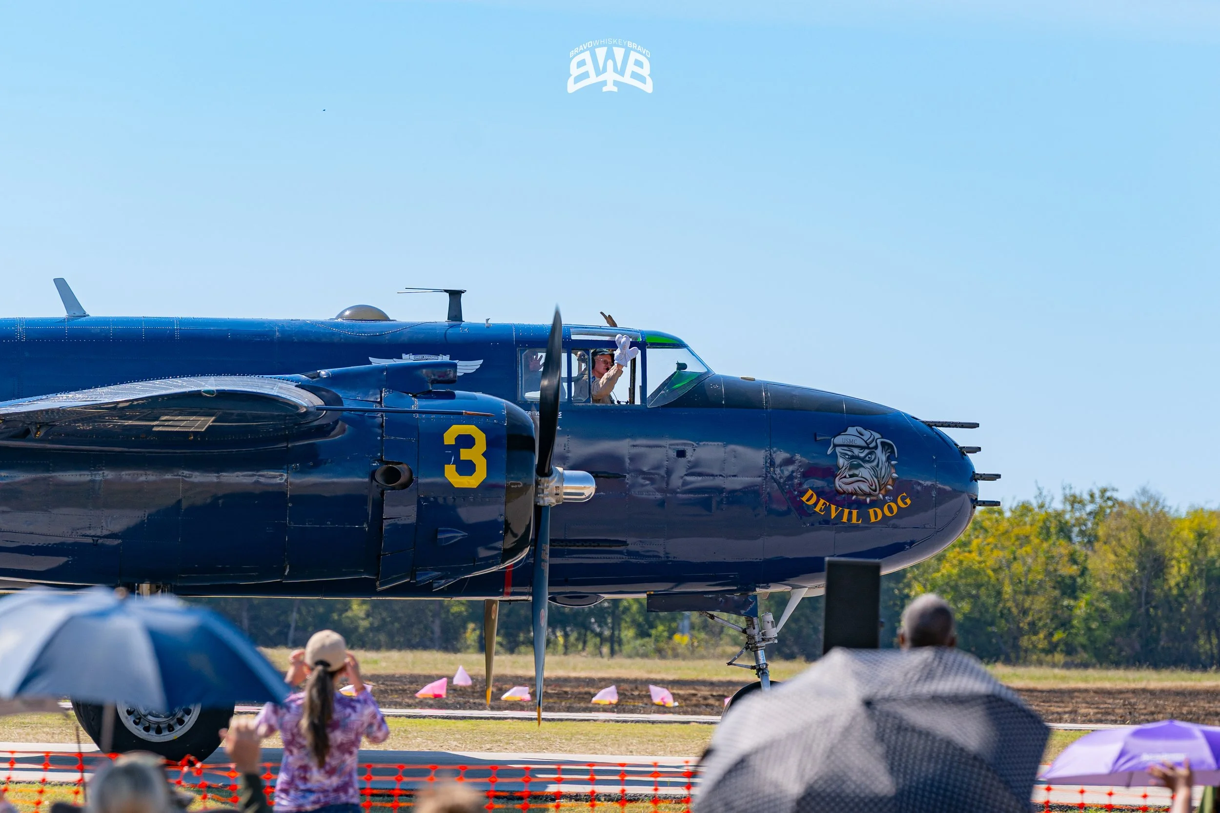 A vintage military aircraft with a dark blue body and the number 3 on its side, featuring a cartoon dog with sunglasses logo that says "Devil Dog," is on display at an airshow. A pilot in the cockpit is waving, while spectators hold umbrellas and wat