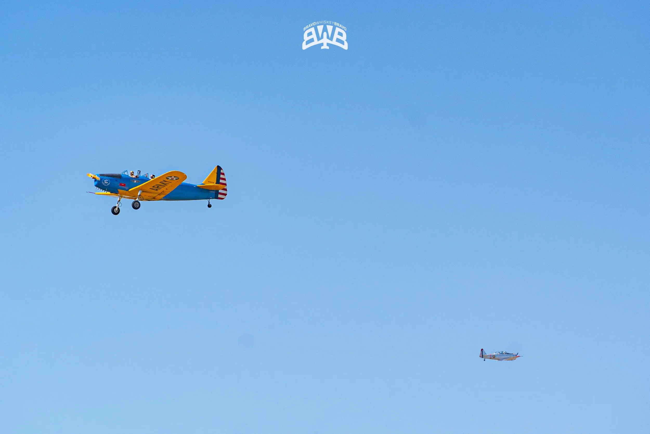 Two small vintage aircraft flying in the sky, one larger plane painted in blue and yellow with American flag colors on the tail, and a smaller plane in silver with red accents.