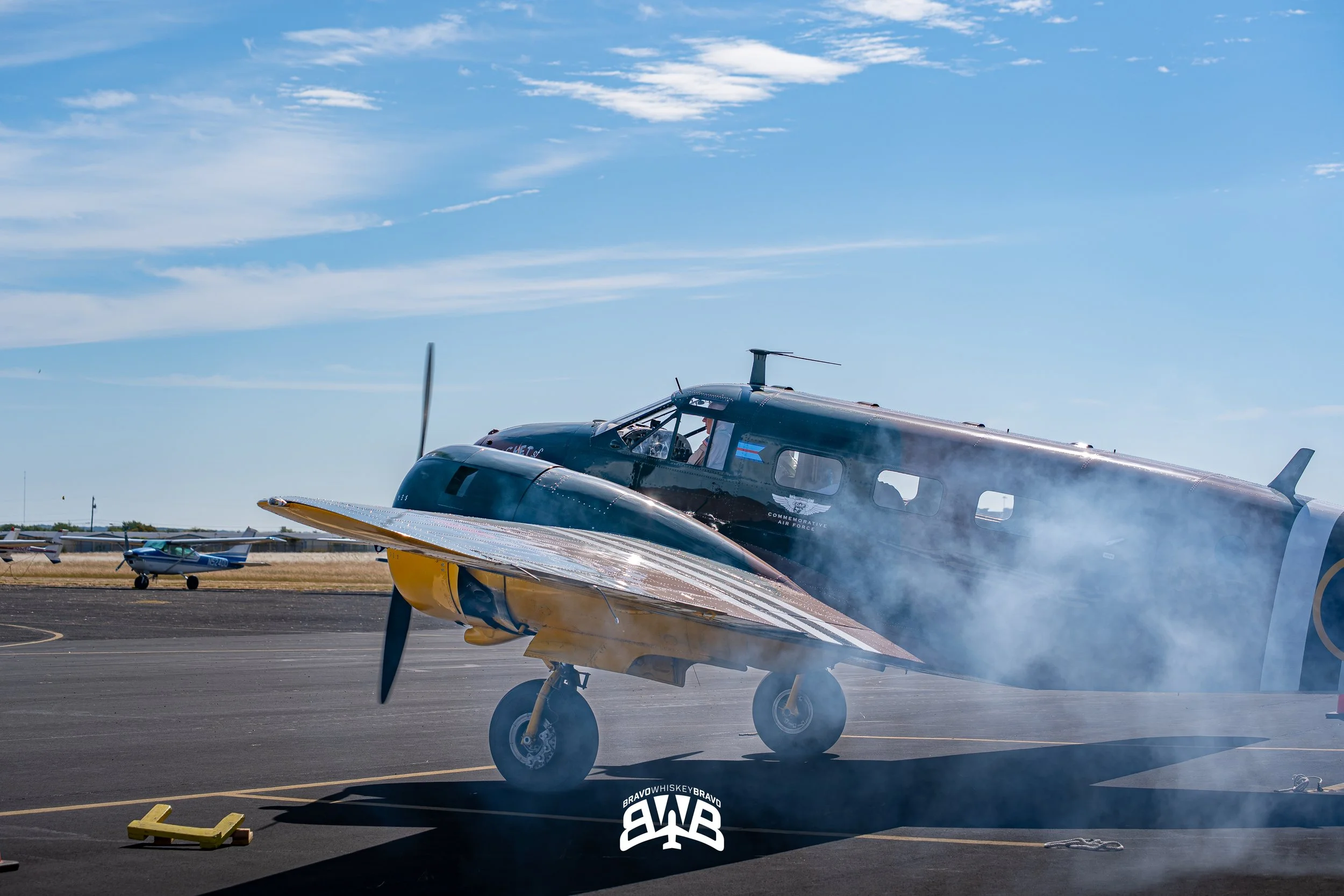 A vintage propeller airplane on the tarmac with smoke around the engine. Smaller airplanes are visible in the background under a partly cloudy sky.