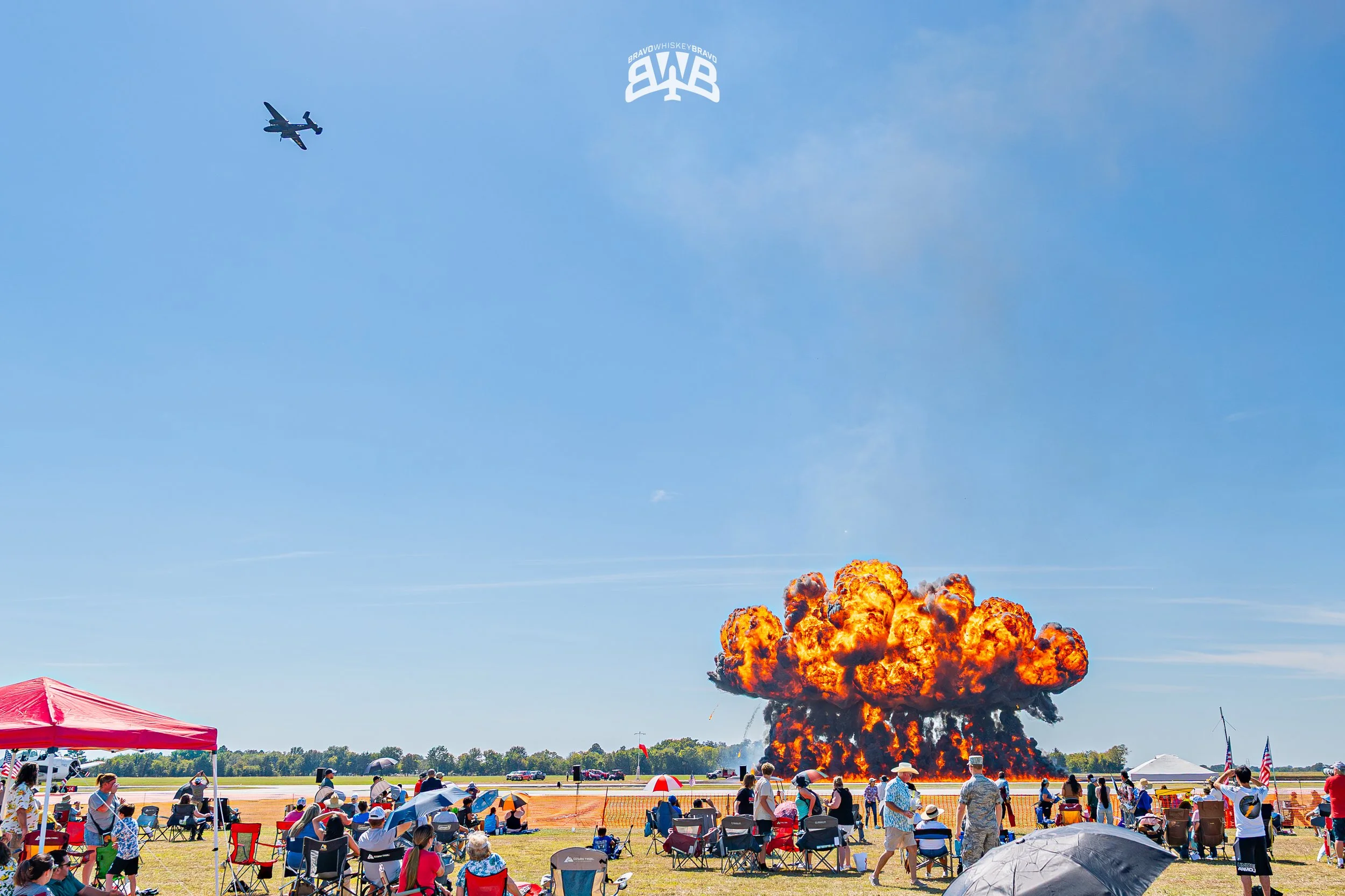 Large explosion on airport runway with a mushroom cloud, spectators watching from the grass, some with umbrellas, tents, and American flags, airplane flying in the blue sky.