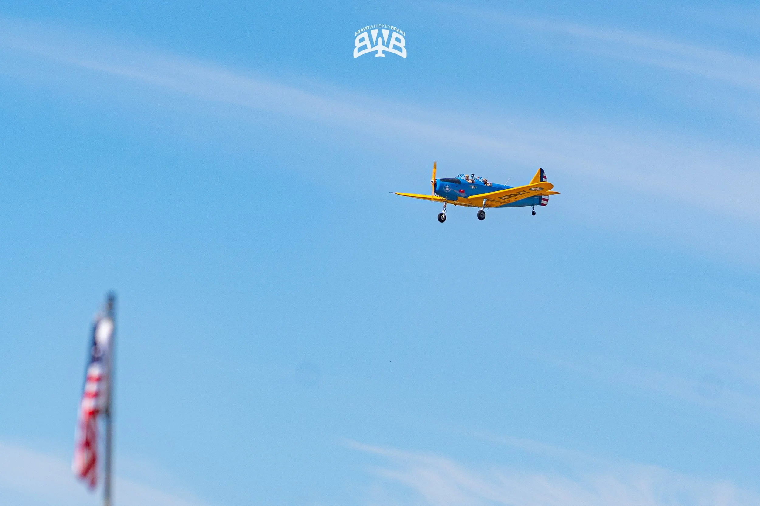 A vintage blue and yellow airplane flying against a clear blue sky, with a blurred American flag in the foreground and an upside-down logo reading 'BRAVO WHISKEY BRAVO' at the top of the image.