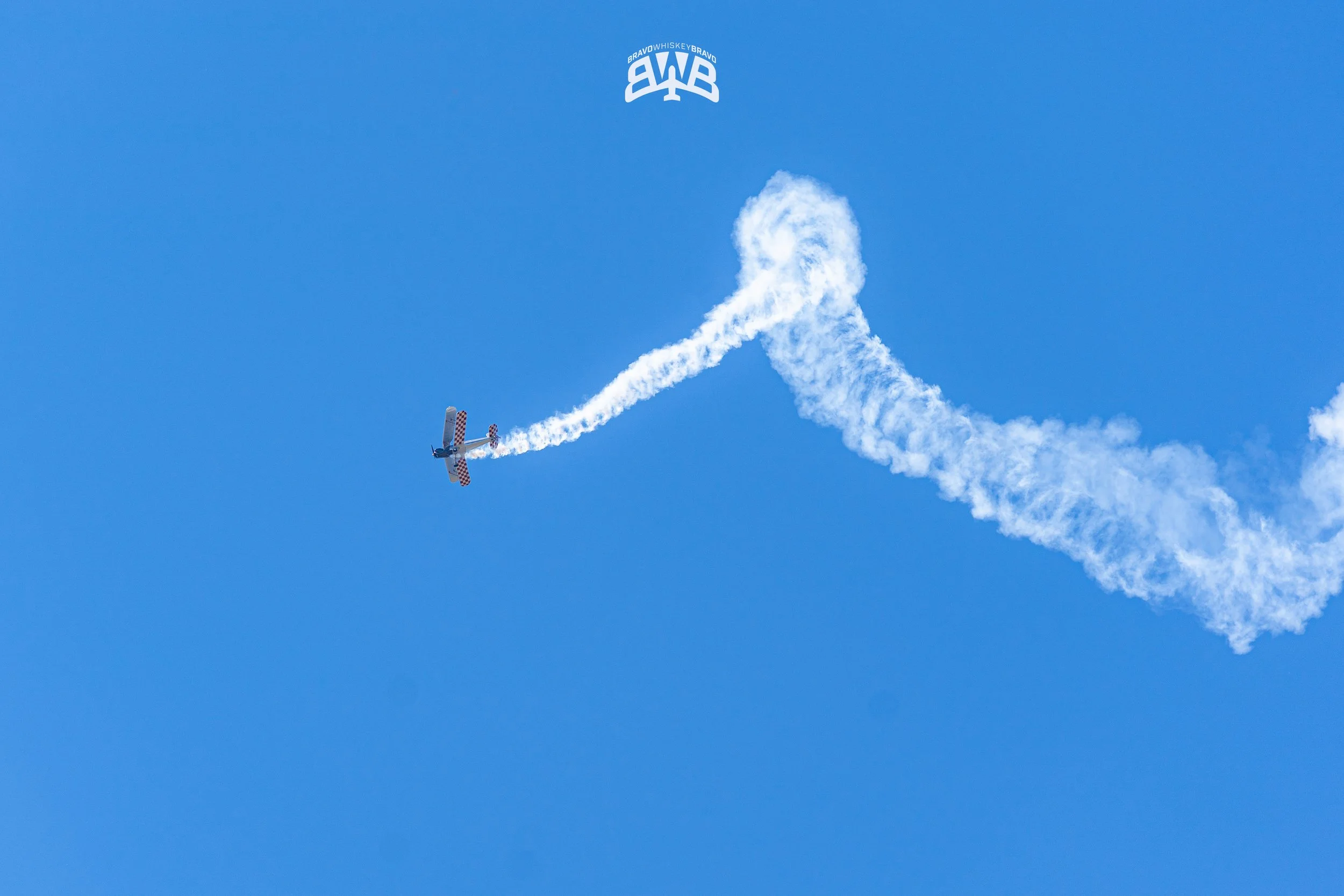 An airplane performing an aerobatic stunt in the sky, leaving a trail of white smoke behind it, with a blue sky background and a small logo at the top reading BWB.