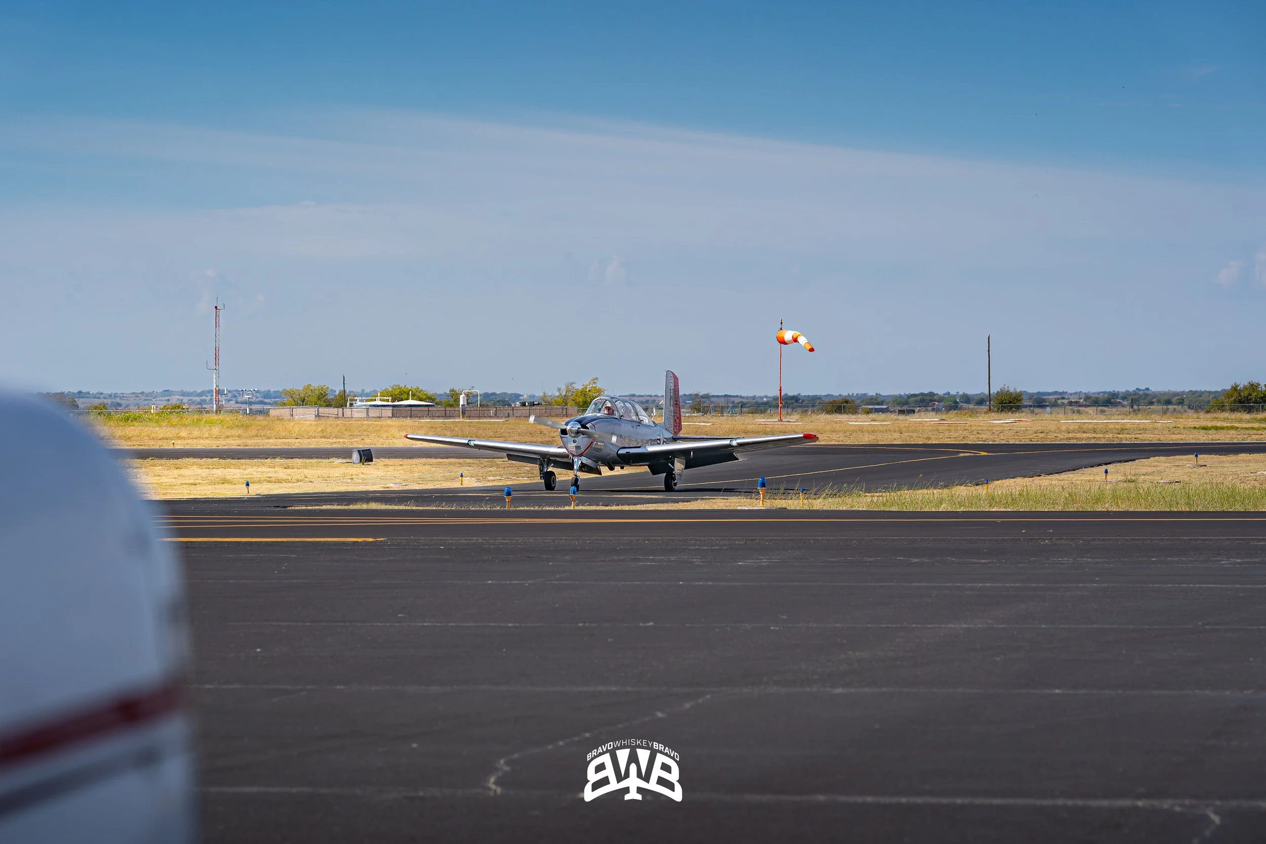 Small vintage airplane on taxiway at an airport, with a windsock and runway markers visible, under a partly cloudy sky.